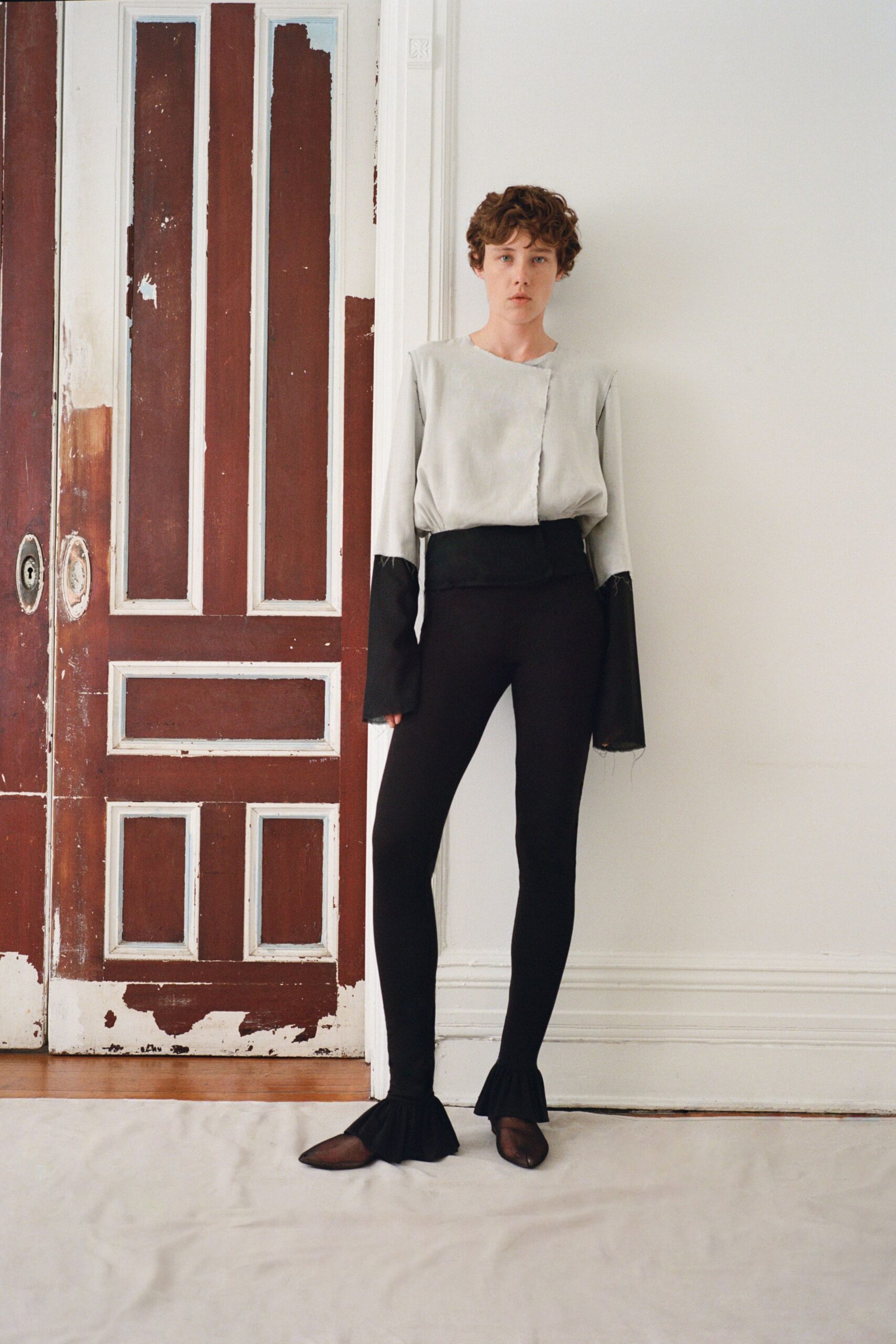 Pale grey satin blouse with an off-center raw-edged closure. Black sheer sleeve extensions hanging past the wrist with loose threads. Opaque black leggings with a wide folded waistband and ruffled hems. Brown mesh flats, pointed and nearly transparent at the toe. In a half-renovated apartment before the movers bring the bed up.