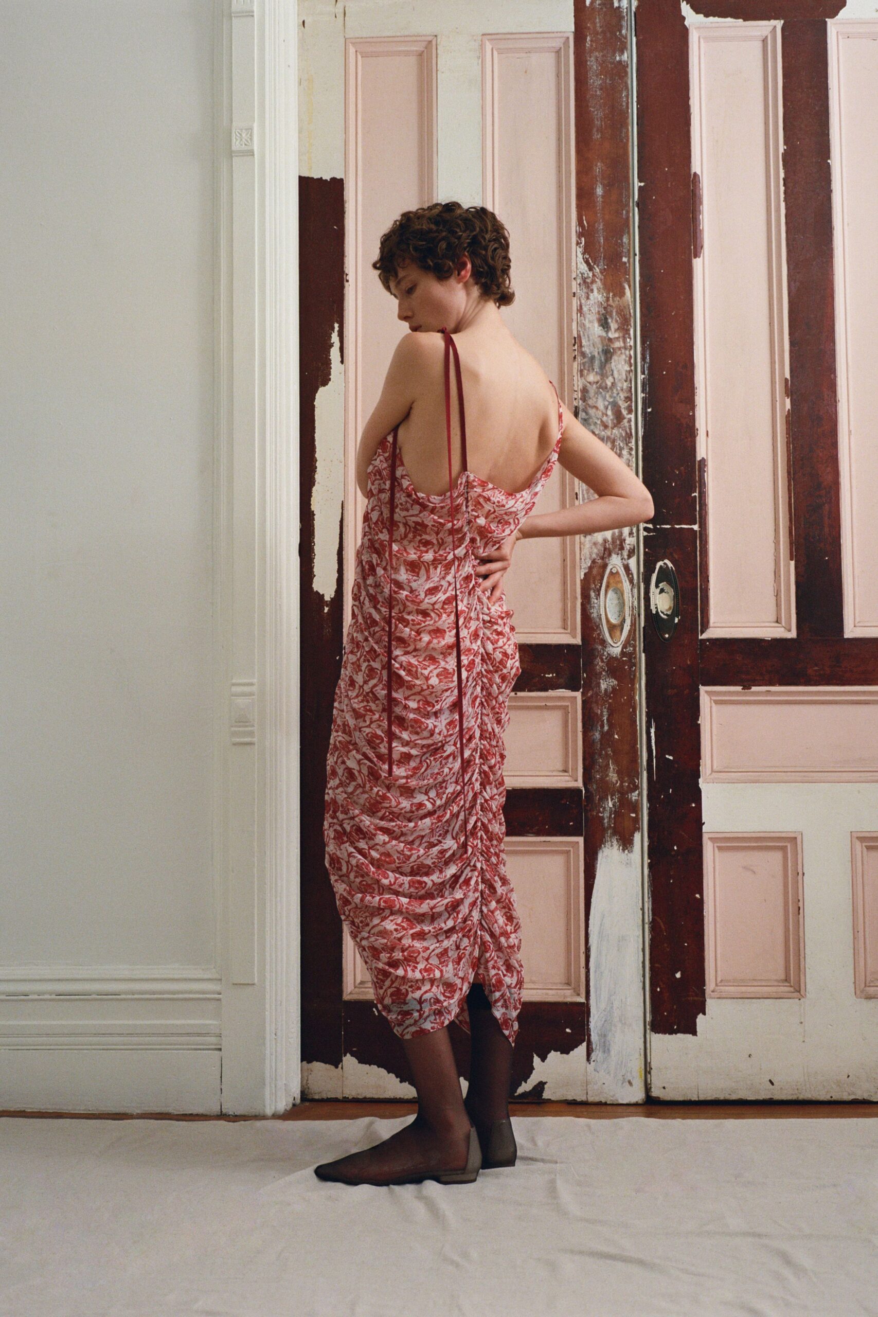 Blush and coral printed slip dress, low cowl back with ruching pulled by long cords. Sheer smoke stockings. Square-toe taupe flats. Afternoon in a half-stripped apartment, paint dust still on the doorframes.