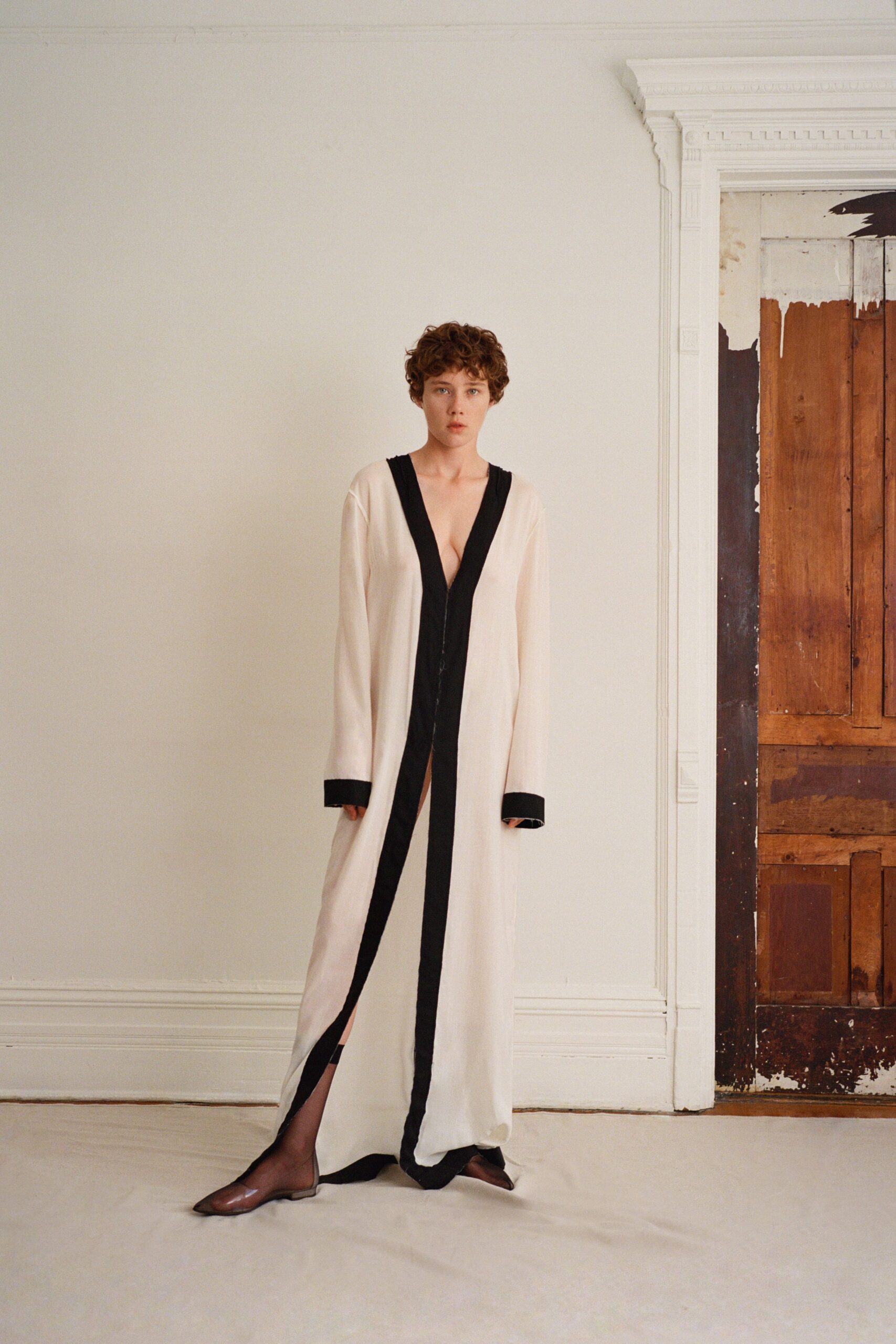 Ivory floor-length dress in fluid crepe, framed by broad black bands and a plunging zip front. Sheer black tights visible through the high front opening. Brown leather pointed flats, low and almost slipper-thin. First cold morning in a rented house with half-stripped doors.