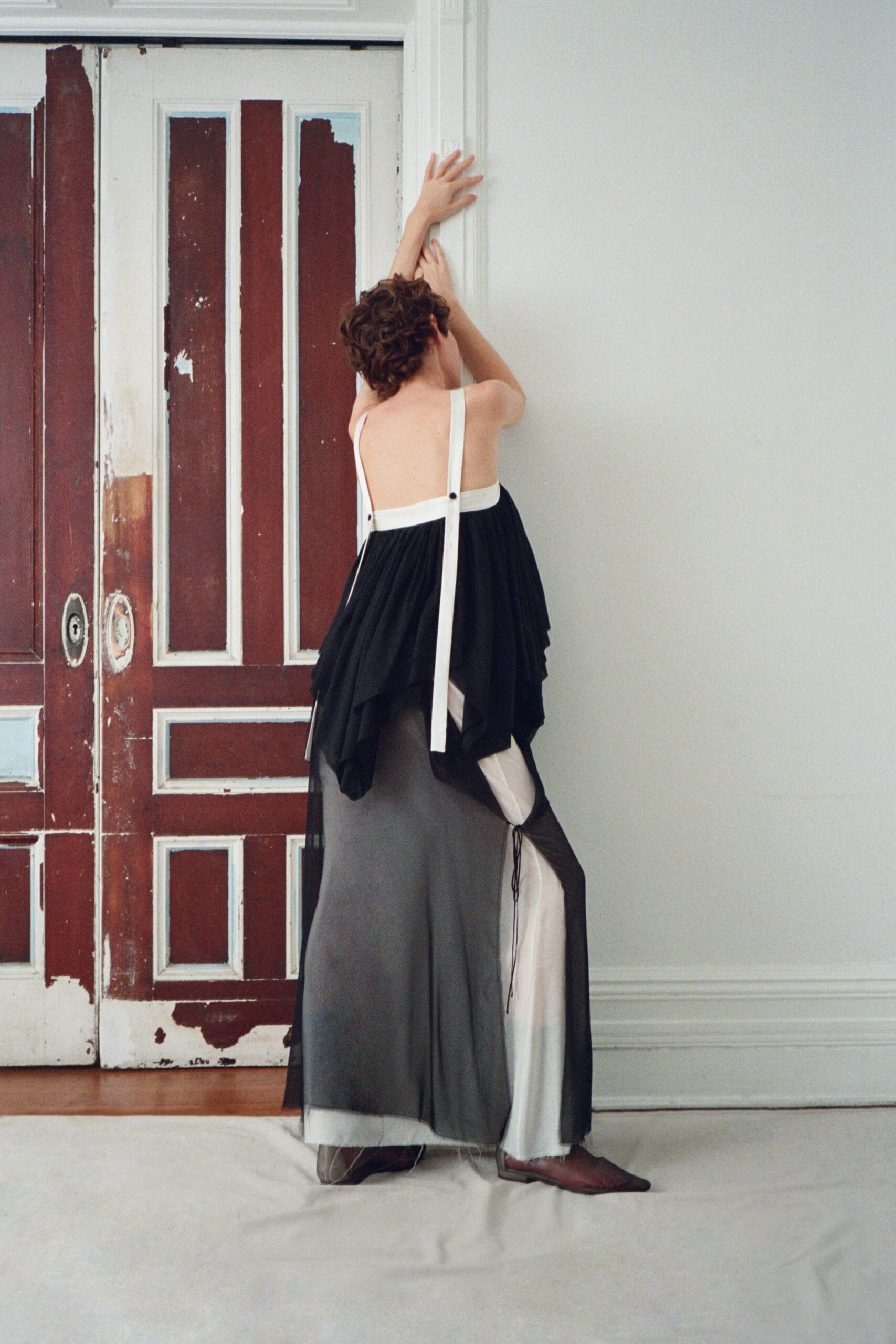 Black chiffon dress with a bare back and long white suspender straps. Ivory satin panel showing through the split side, tied with a loose cord. Pointed brown flats, almost hidden under the sheer hem. In a rented mansion while the paint on the doors is still peeling.