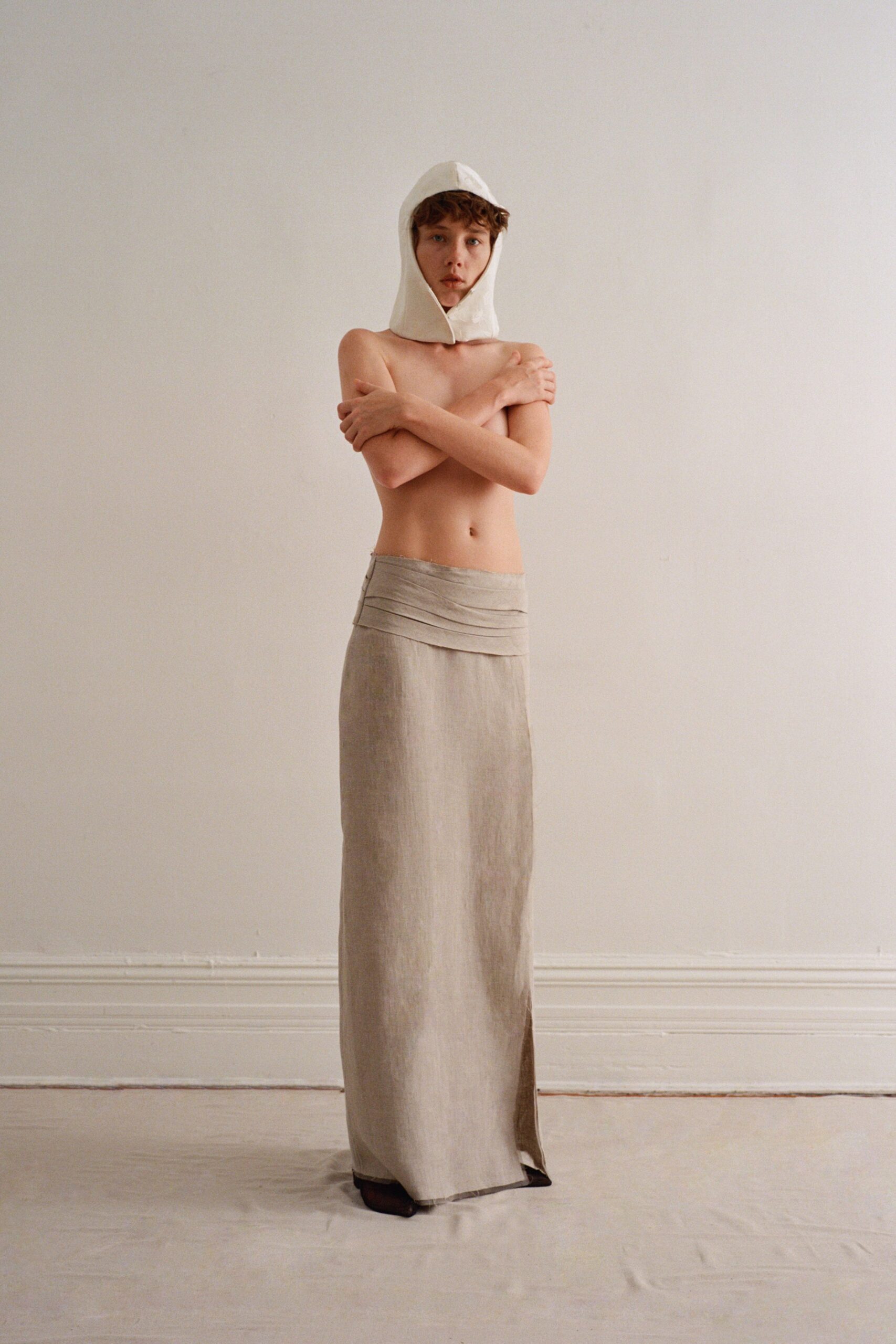 White structured hood with a pointed throat opening framing the face. Long oat linen skirt, folded waistband wrapping low across the hips. Dark square toes barely breaking the hem. Bare apartment floors on the first warm day you open every window.
