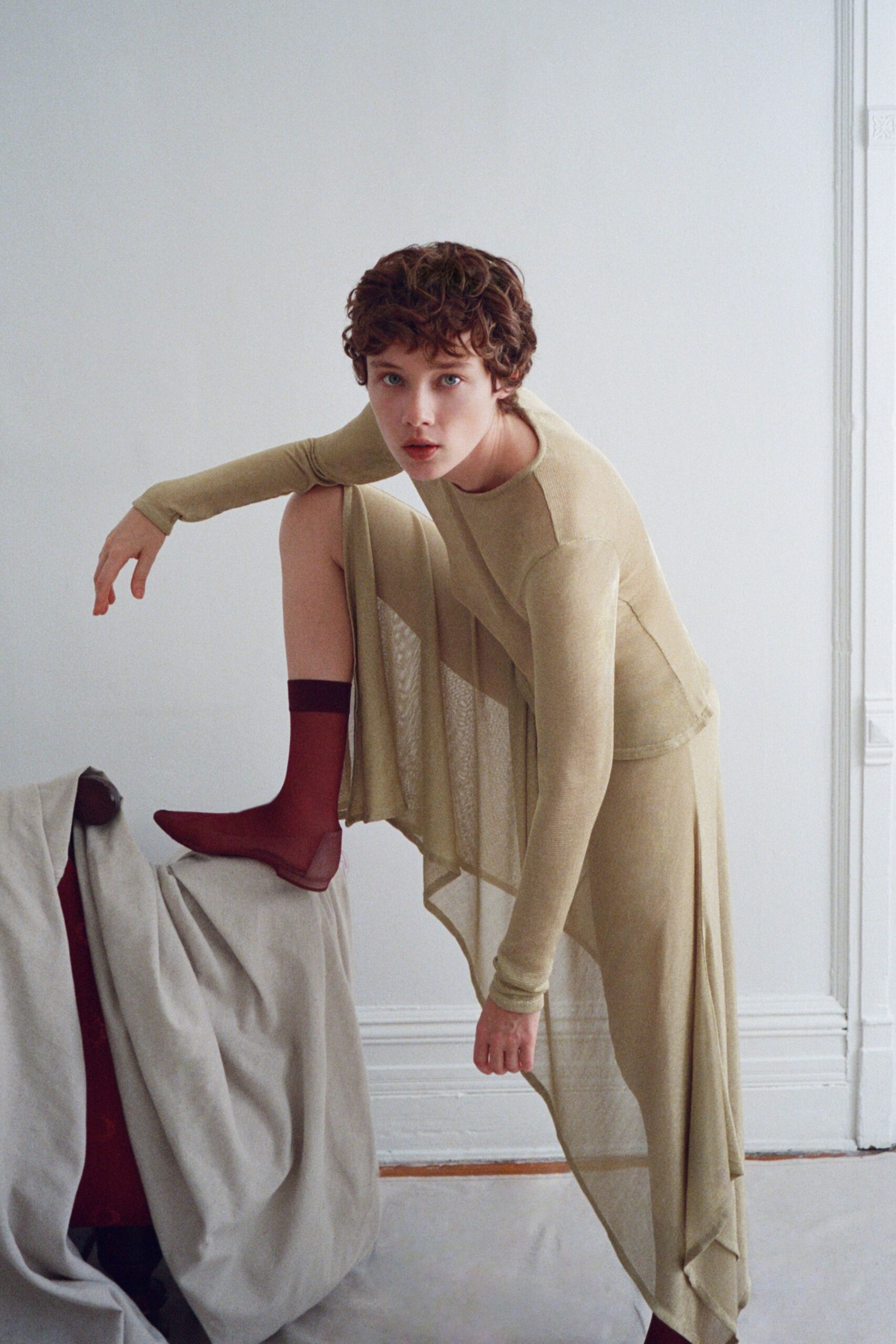 Moss-beige fine knit top with long sleeves and a raw curved seam. Sheer panelled overskirt falling in uneven handkerchief points. Matching close knit leggings under the drape. Oxblood sheer socks with a dark band at the calf. Crossing a cold studio floor before the radiators wake up.
