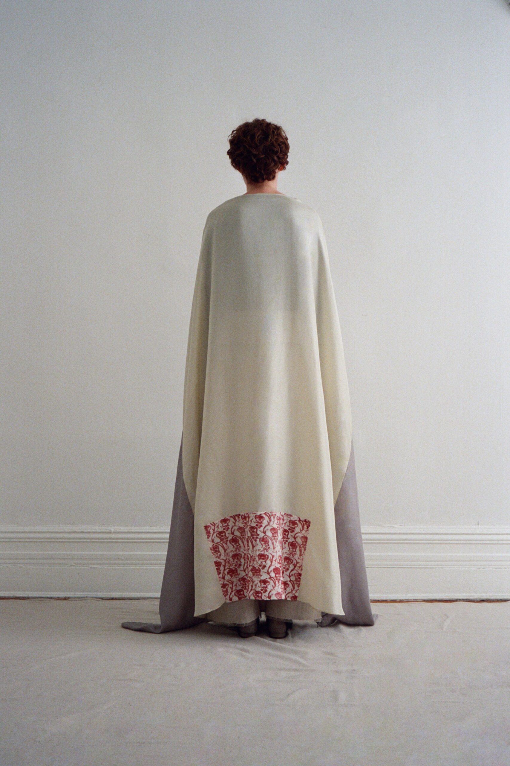 Ivory floor-length cape in heavy felted wool, red block-print panel at the hem. Pale dove underlayer spreading out in a soft train behind the cape. Square-toed shoes barely showing. In the hush after a small civil ceremony, facing the empty room.