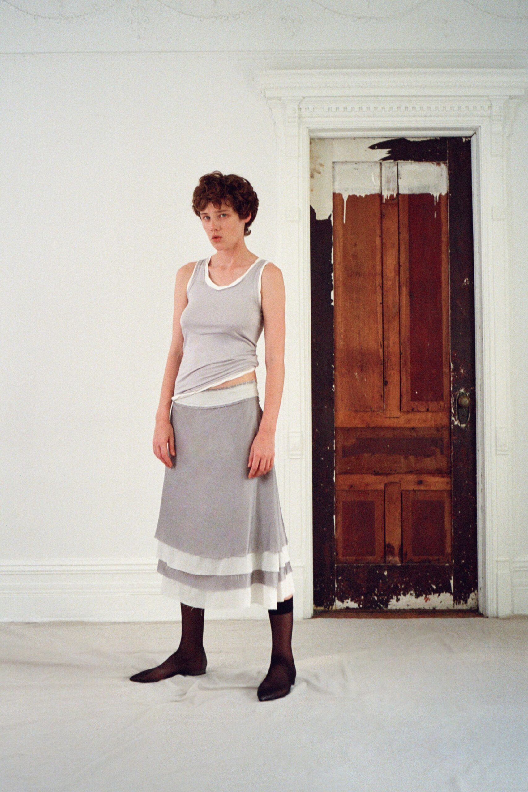 Pale grey jersey tank, one side twisted up to expose the underlayer. Misty grey skirt with sheer organza bands stacked near the hem. Black sheer stockings. Pointed black flats, almost socklike. Sunday in a rented house, paint still drying on the bedroom door.