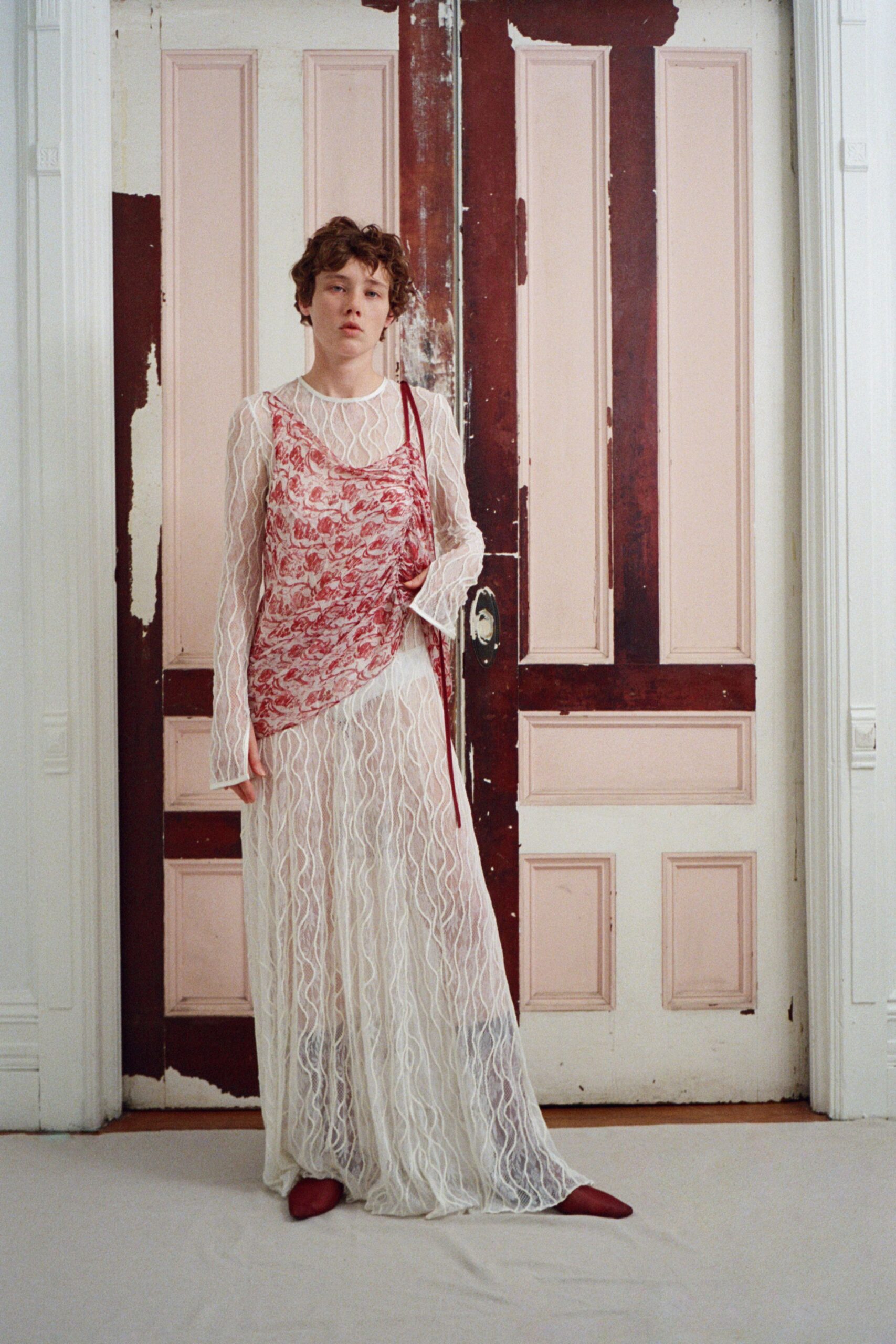 Sheer white mesh dress stitched all over with squiggled cord lines. Red marbled slip hanging askew, one long ribbon strap falling loose. Pointed oxblood boots peeking bluntly from the hem. Opening the locked room in a faded seaside guesthouse at noon.