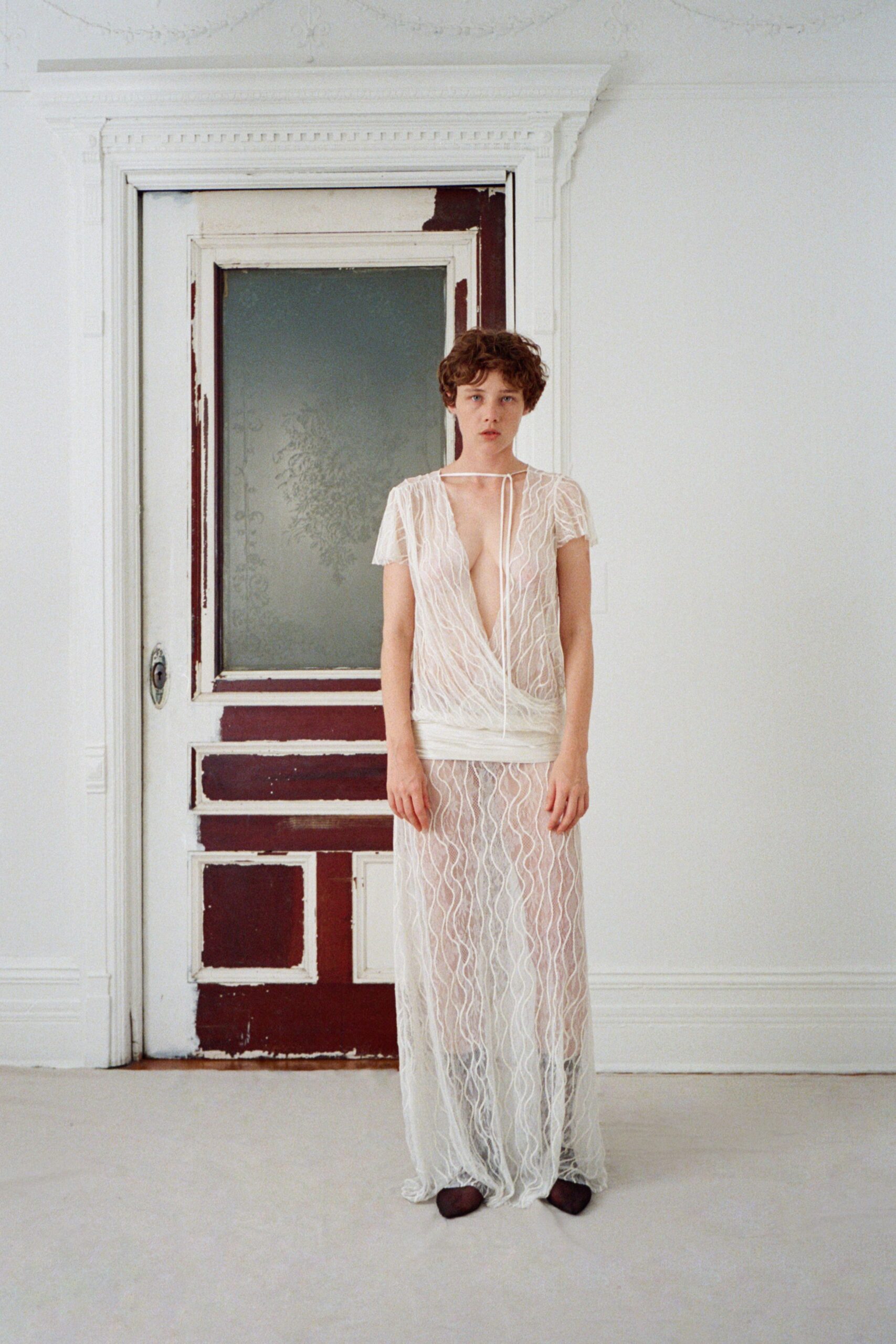 Sheer ivory mesh top traced with raised squiggle embroidery, plunging to the waist. Long self tie looped once at the collarbone. Matching column skirt in the same wavy embroidery, gauze band hugging low hips. Dark pointed flats under the transparent hem. Morning in a rented villa, paint flaking from the doors.