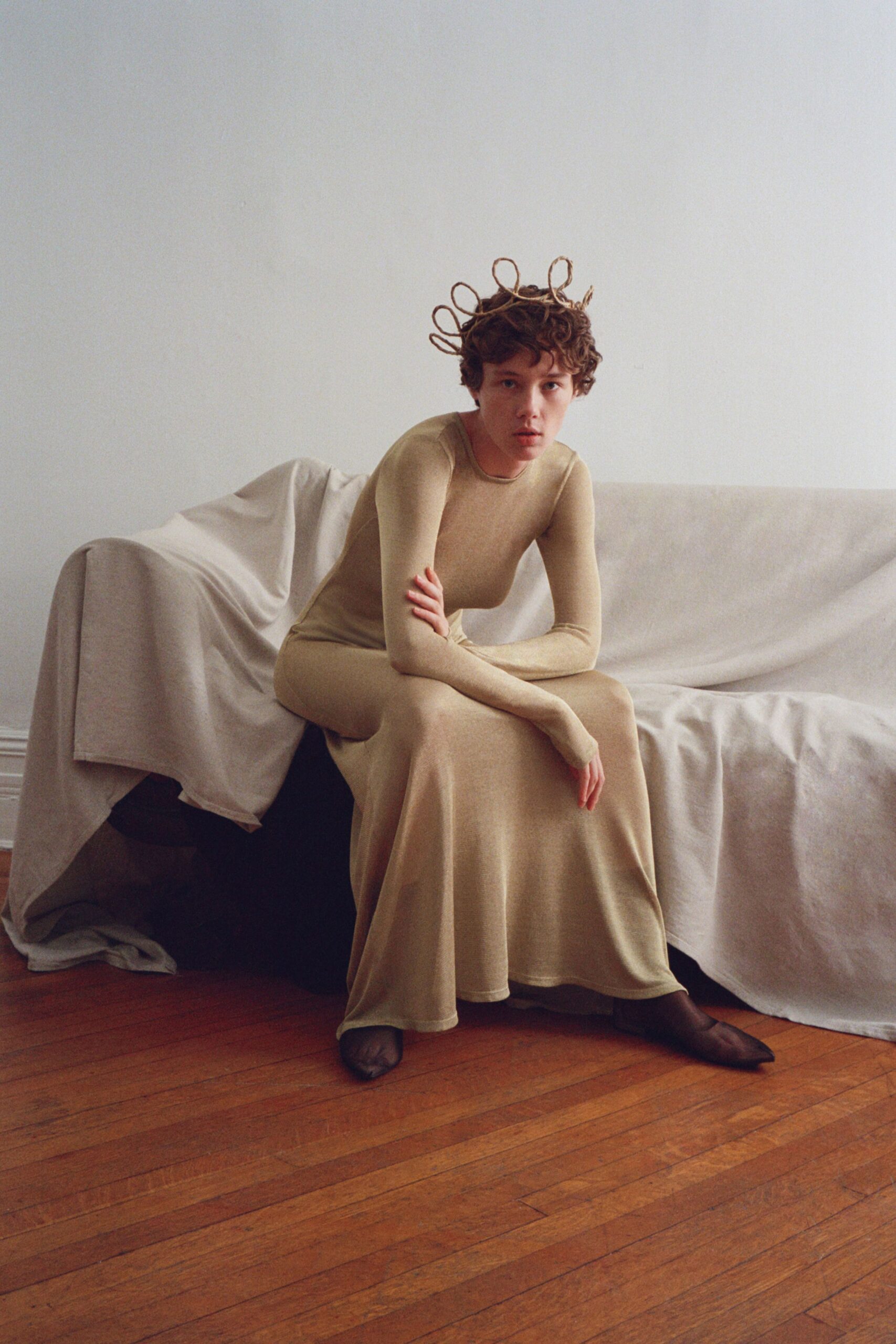 Looped twig crown, each bent branch rising in uneven halos. Pale sand jersey dress, cut close through the arms then pooling at the hem. Sheer black stockings, toe seams visible against the floor. Sitting very still after bringing branches in from the cold room.