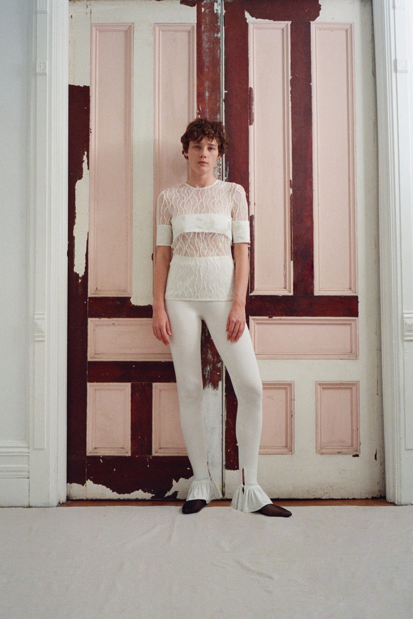 Sheer ivory mesh top traced with wavy stitched channels and satin bands. Opaque ivory leggings ending in gathered ruffle hems at the ankle. Dark almond-toe flats. Morning in a half-painted townhouse, dust sheets still taped to the floor.