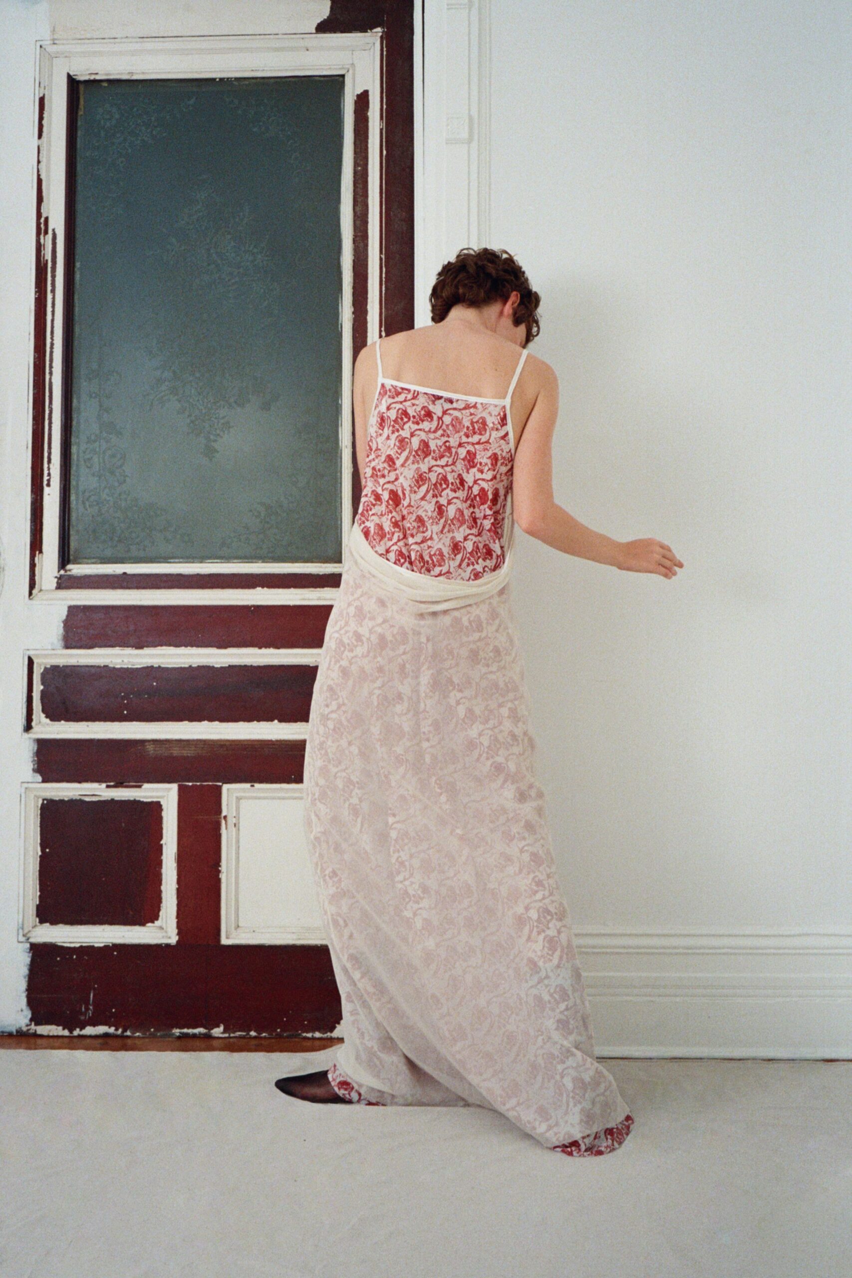 Red toile slip dress with narrow straps and a low square back. Sheer blush overskirt in gauze, knotted low across the hips. Black pointed flats peeking under the dragging printed hem. Crossing a rented apartment in stocking feet, windows cracked for the heat.
