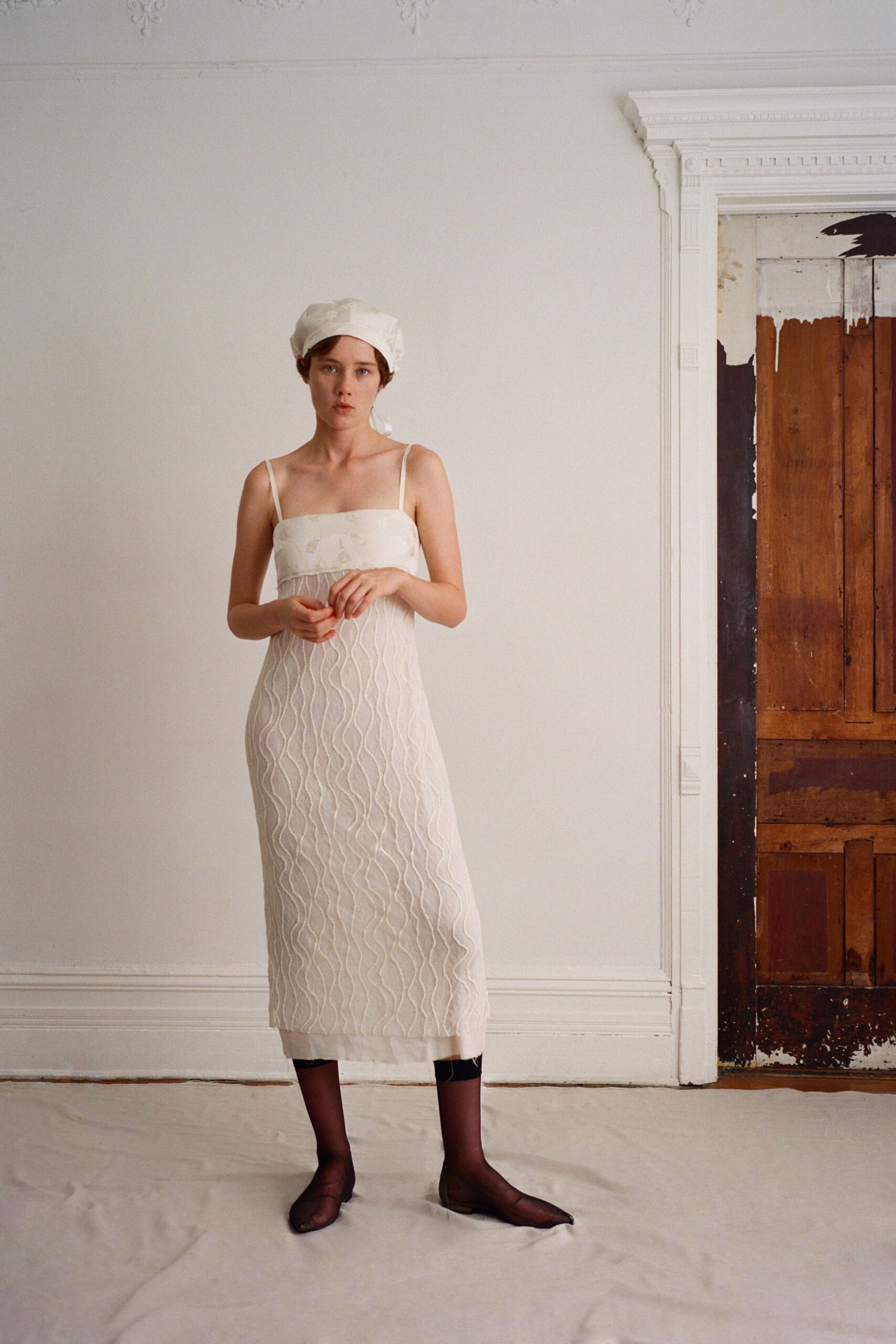 White cotton headscarf tied low, one tail peeking behind the ear. Ivory midi slip dress with narrow straps and raised squiggle quilting. Sheer black stockings ending abruptly below the calf. Brown mesh flats with a blunt squared toe. In a rented room before the painter comes back for the ladders.