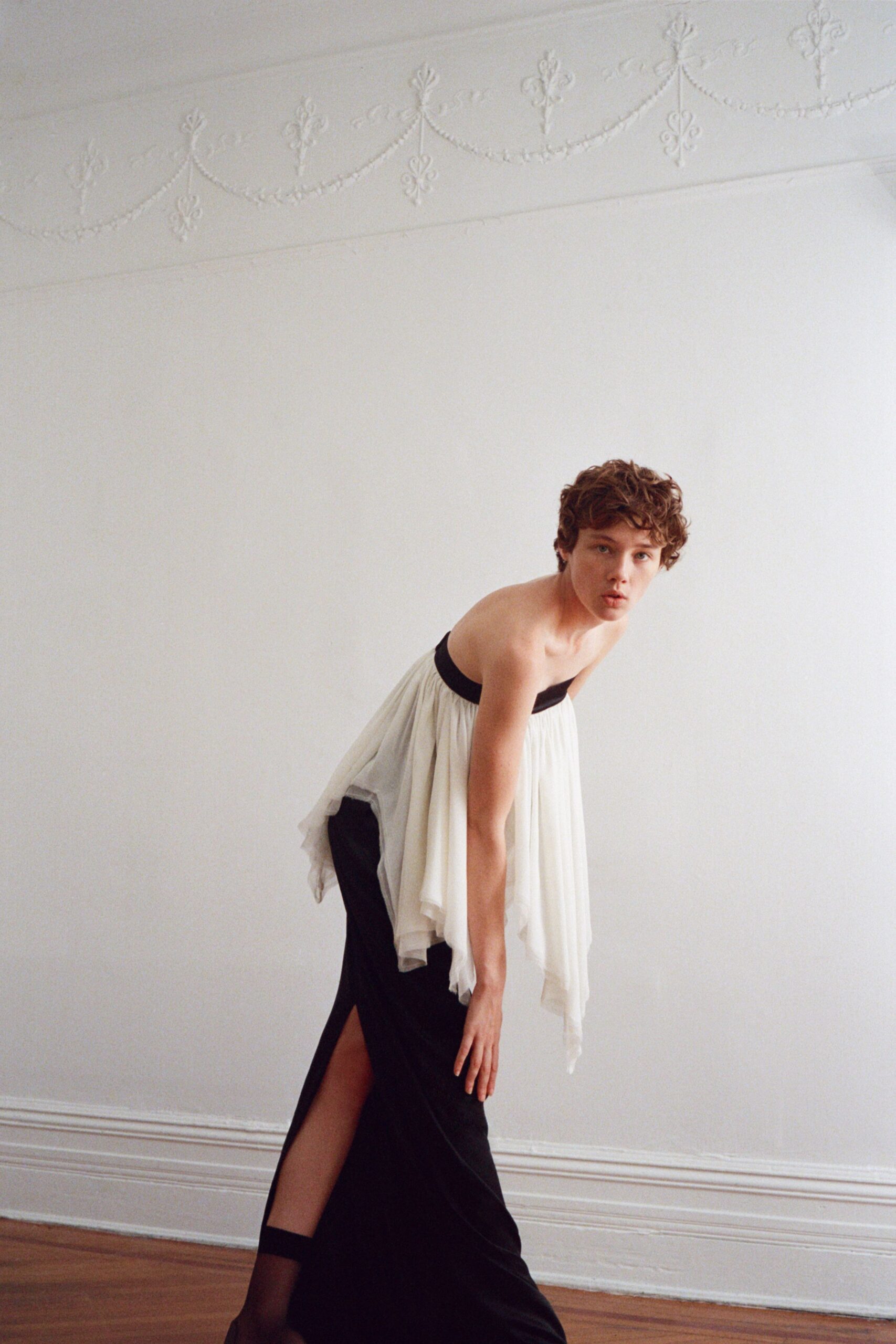 Strapless white chiffon overdress, gathered to a narrow black band. Black column skirt with a thigh slit opening under the drape. Sheer black thigh-highs with a dark stay band. In a rented parlour before the windows are opened for summer.