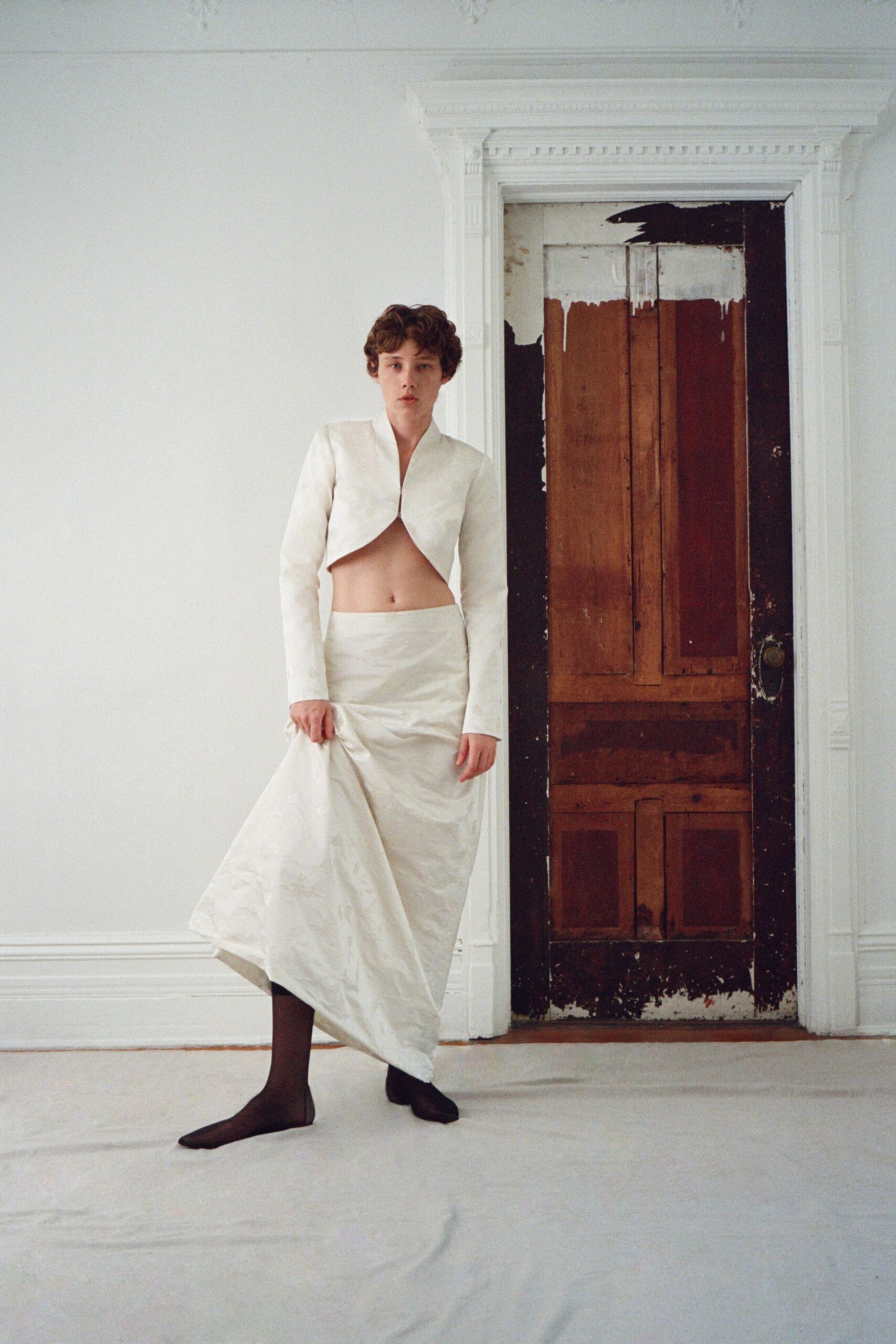 Ivory cropped jacket cut away beneath the bust, collarless and close. Matching satin column skirt, one side hitched up in the model's fist. Sheer black tights. Square-toe black flats. The courthouse is closed, but you still have the flowers in the car.
