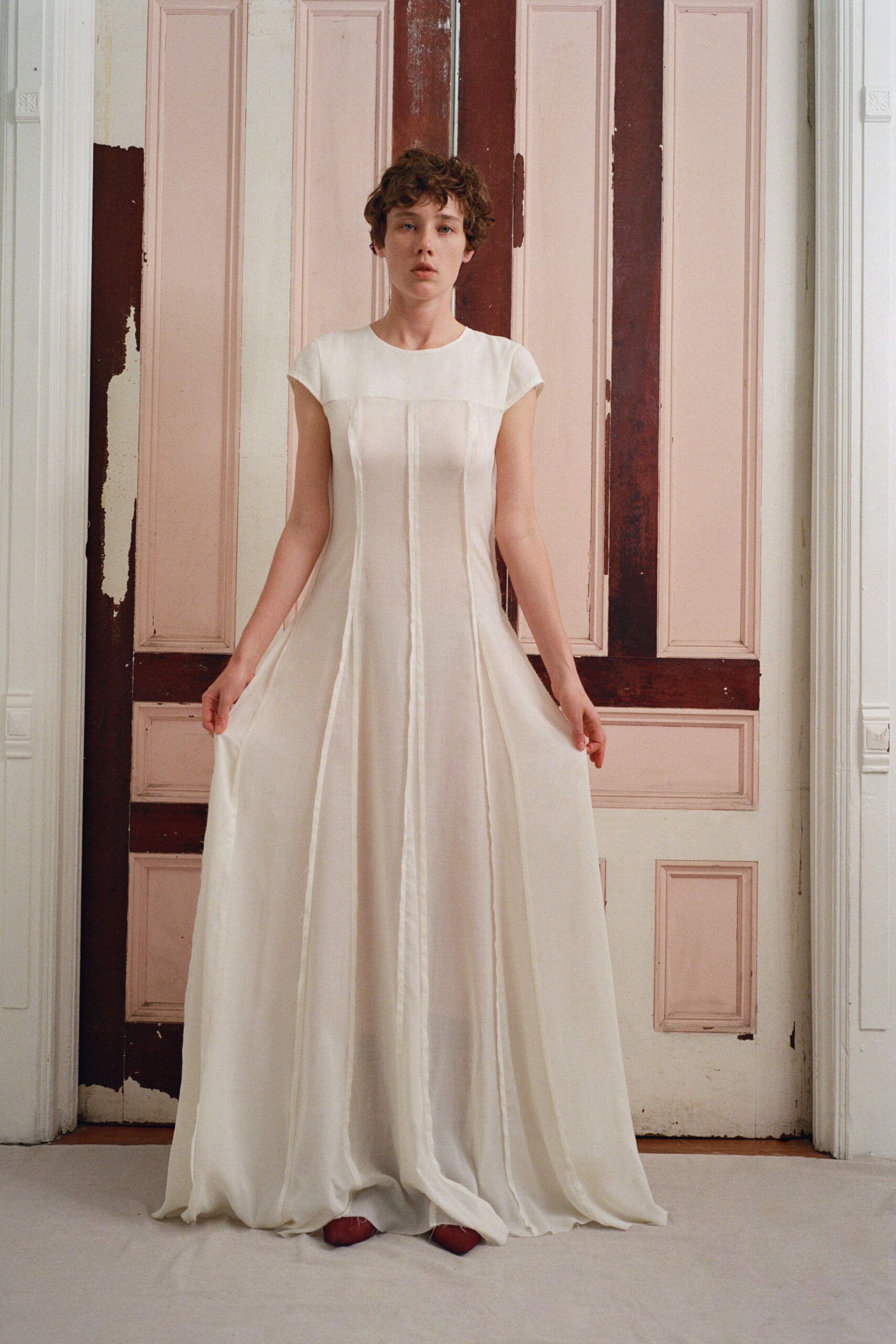 Milk-white floor-length dress with cap sleeves and raised seam channels down the skirt. Rust-red pointed shoes peeking from the pooled hem. In a faded ballroom before the guests arrive and the windows are opened.