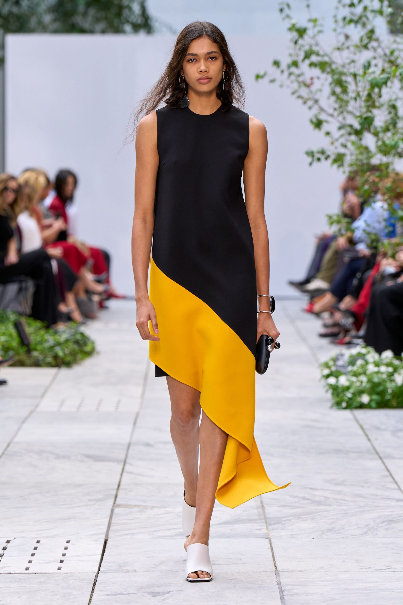 Black shift dress cut in dense crepe, sliced by a marigold draped panel. Long black drop earrings. Silver cuff with a dark oval inset. Black hard clutch with pearl-like ball ends. White leather mules, squared and open at the toe. Crossing a sculpture court at noon, carrying nothing but your keys.