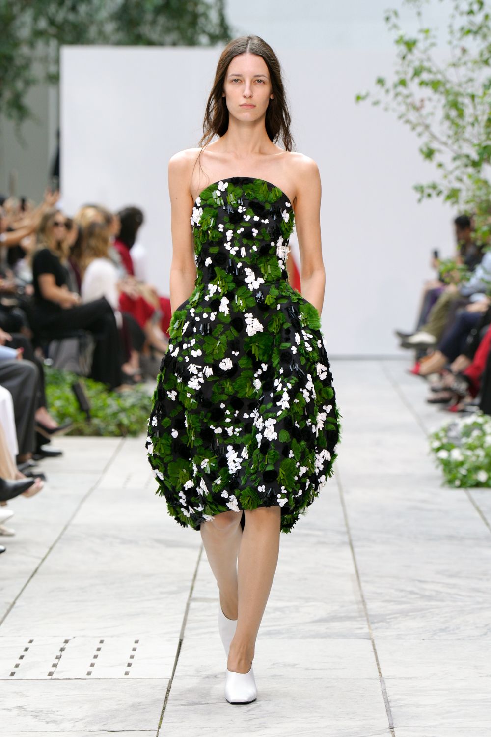 Strapless black cocktail dress densely appliqued with green leaves and white blossoms, skirt puffed into rounded panniers. White square-toe pumps disappearing under the hem. Garden wedding reception, after the speeches, crossing the lawn for air.
