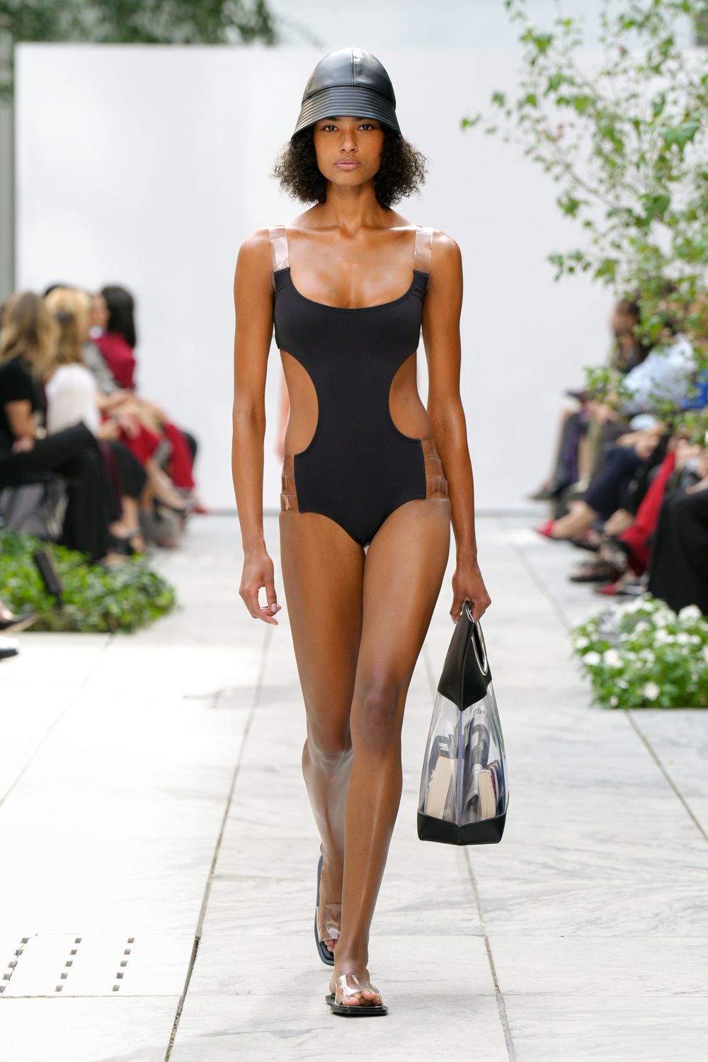 Black leather cap with tight concentric topstitching across the brim. Black maillot cut deep at the sides, held by clear vinyl tabs. Transparent tote with black leather corners and rolled handles. Flat black sandals with cloudy PVC straps over the toes. Pool deck before noon, hotel key card warming in your hand.