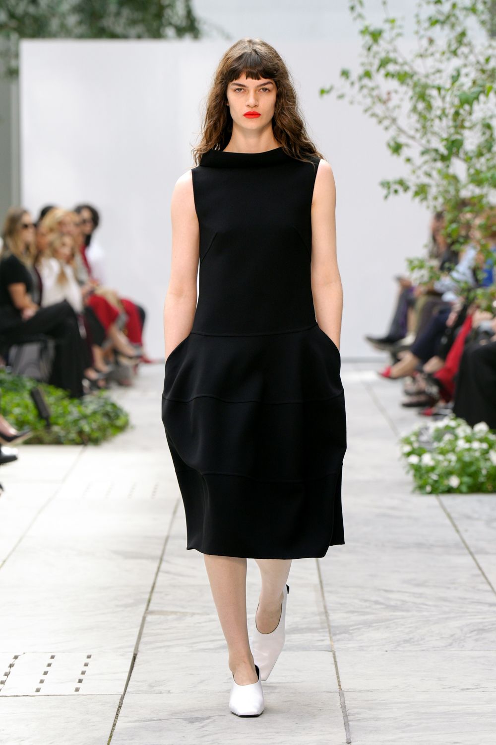 Black sleeveless dress with a rolled bateau neck and tiered cocoon skirt. White leather pumps with sharply squared toes. Crossing a sculpture garden after rain, hands buried in the dress pockets.