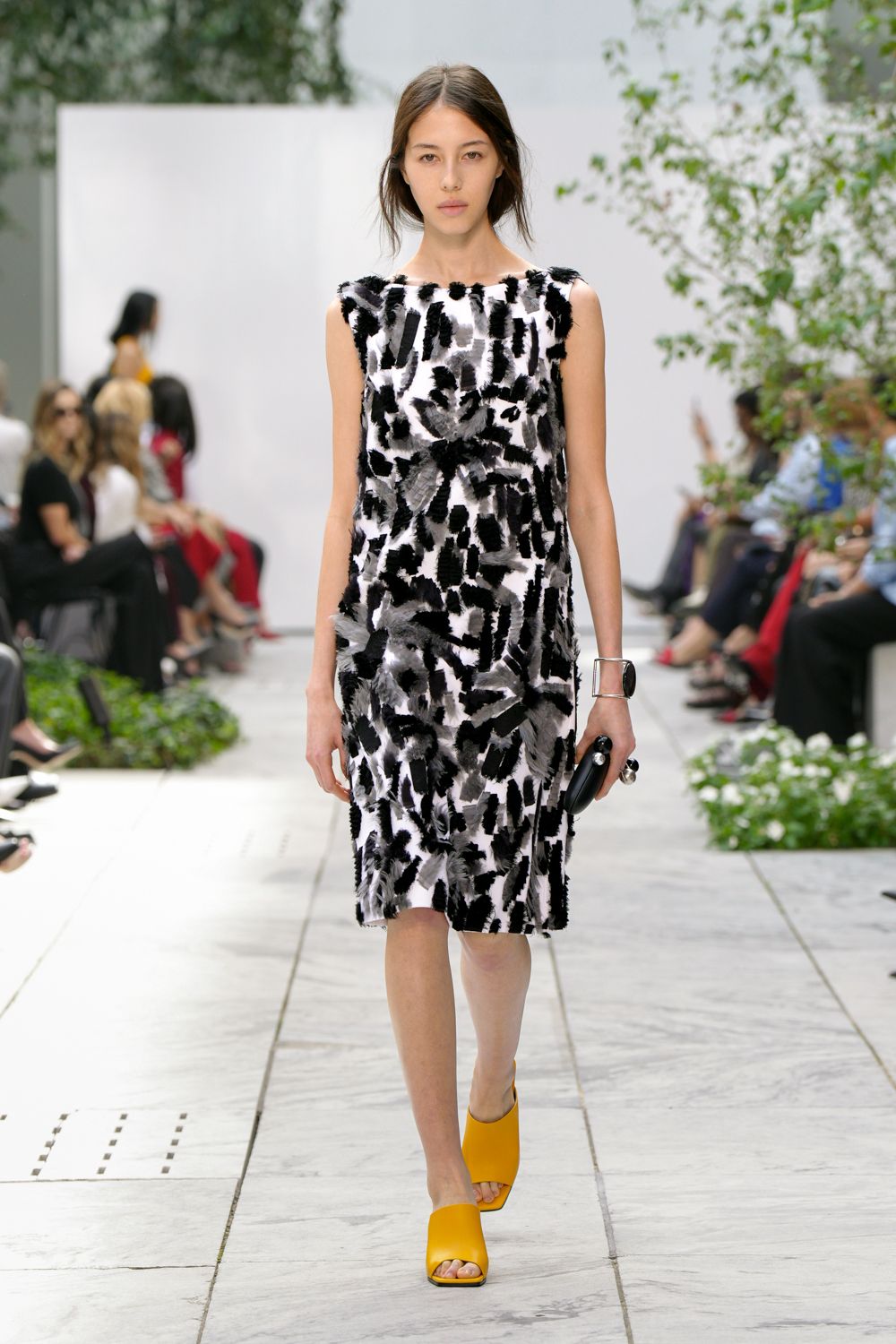 Sleeveless white shift dress densely appliqued with black and dove brushstroke tufts. Wide silver cuff framing a glossy black oval stone. Small black clutch tucked lengthwise in the hand. Marigold leather mules with a square open toe. Crossing a museum courtyard just after the sprinkler mist lifts.