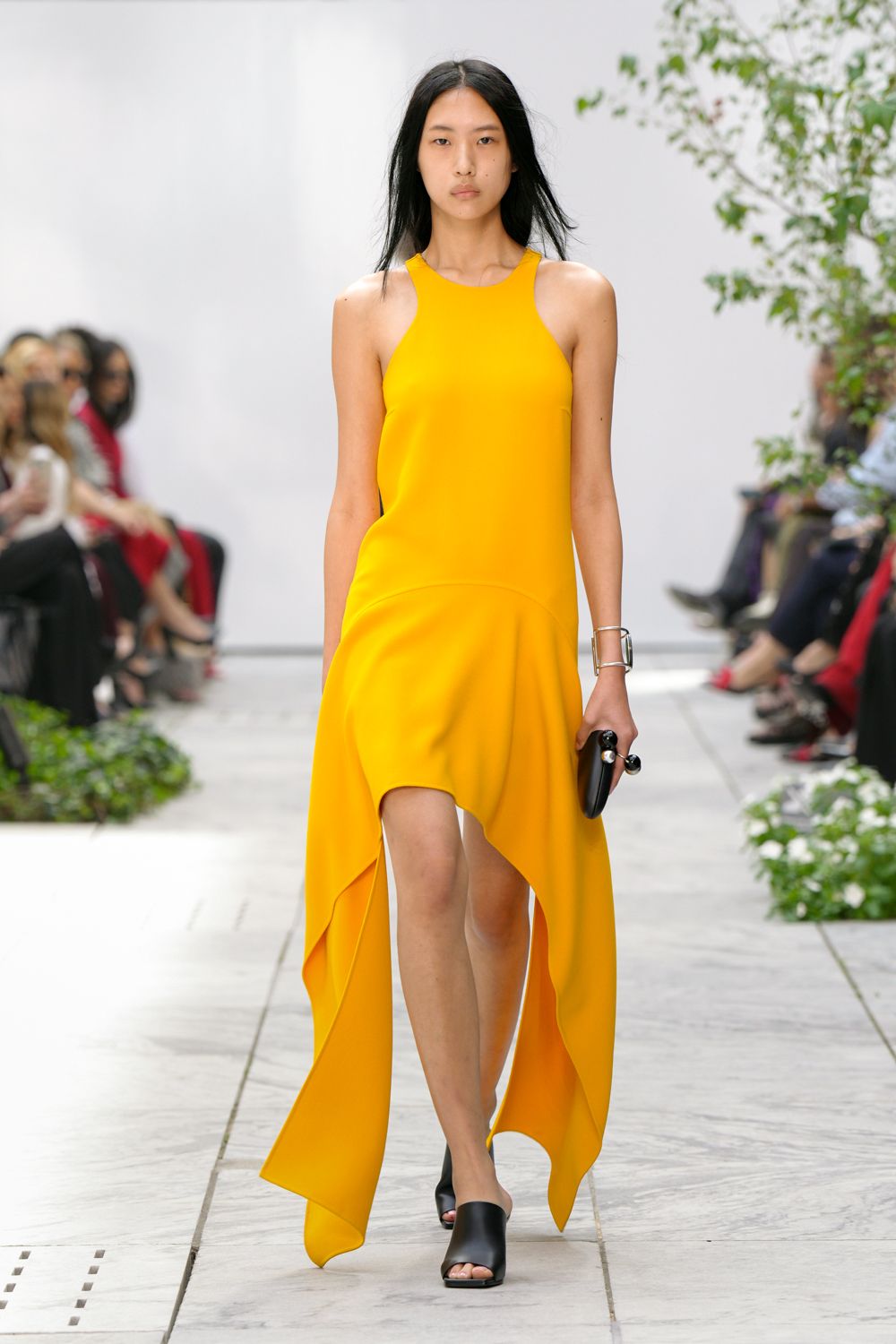 Marigold crepe dress with a high-cut racer front and cascading handkerchief hem. Wide silver cuff, squared and spare at the wrist. Black oval clutch gripped through two glossy ring handles. Black leather mules with a clean open toe. Crossing a sculpture garden just before the caterers clear the glasses.
