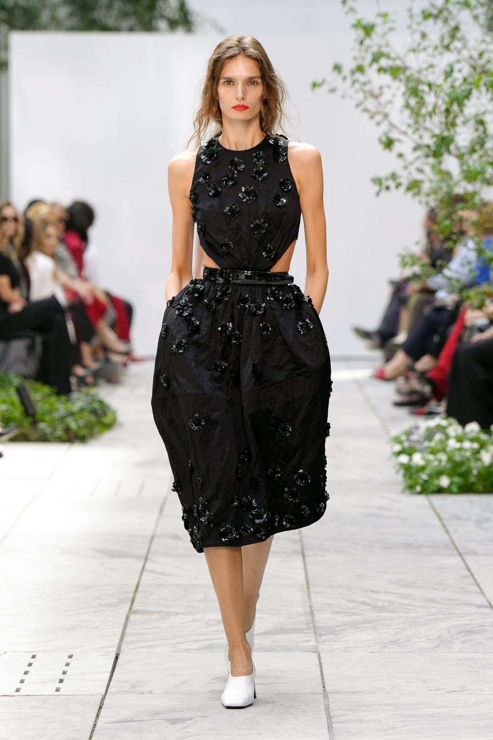Black taffeta dress with side cutouts and scattered lacquered rosettes. Glossy black belt cinching the gathered waist. White leather ankle boots with squared toes. Crossing a courtyard wedding lawn before the speeches start.