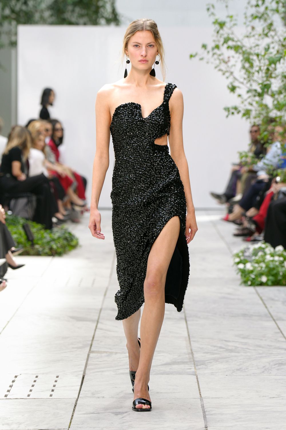 Black drop earrings with two mismatched pebble-like pendants. Black one-shoulder dress densely crusted in glossy paillettes, side cutout and thigh-high split. Black patent peep-toe mules. Garden wedding after midnight, carrying your heels back across the stone path.