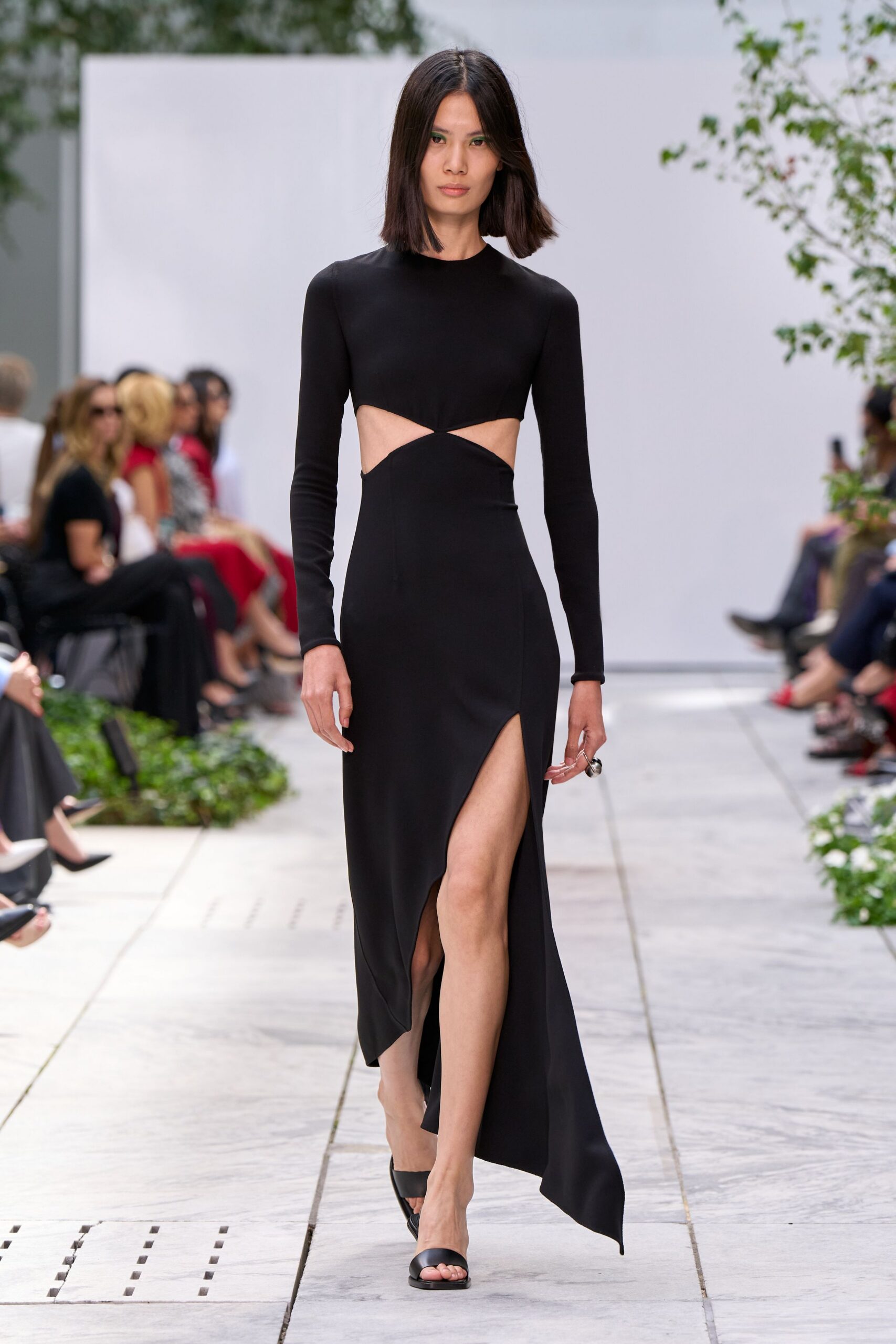 Black jersey column dress with waist cutouts pinched into a sharp twist. Black leather thong sandals with a broad flat toe band. Oversized silver dome ring catching the hand at her side. Walking across a museum courtyard just after the paving stones stop radiating heat.