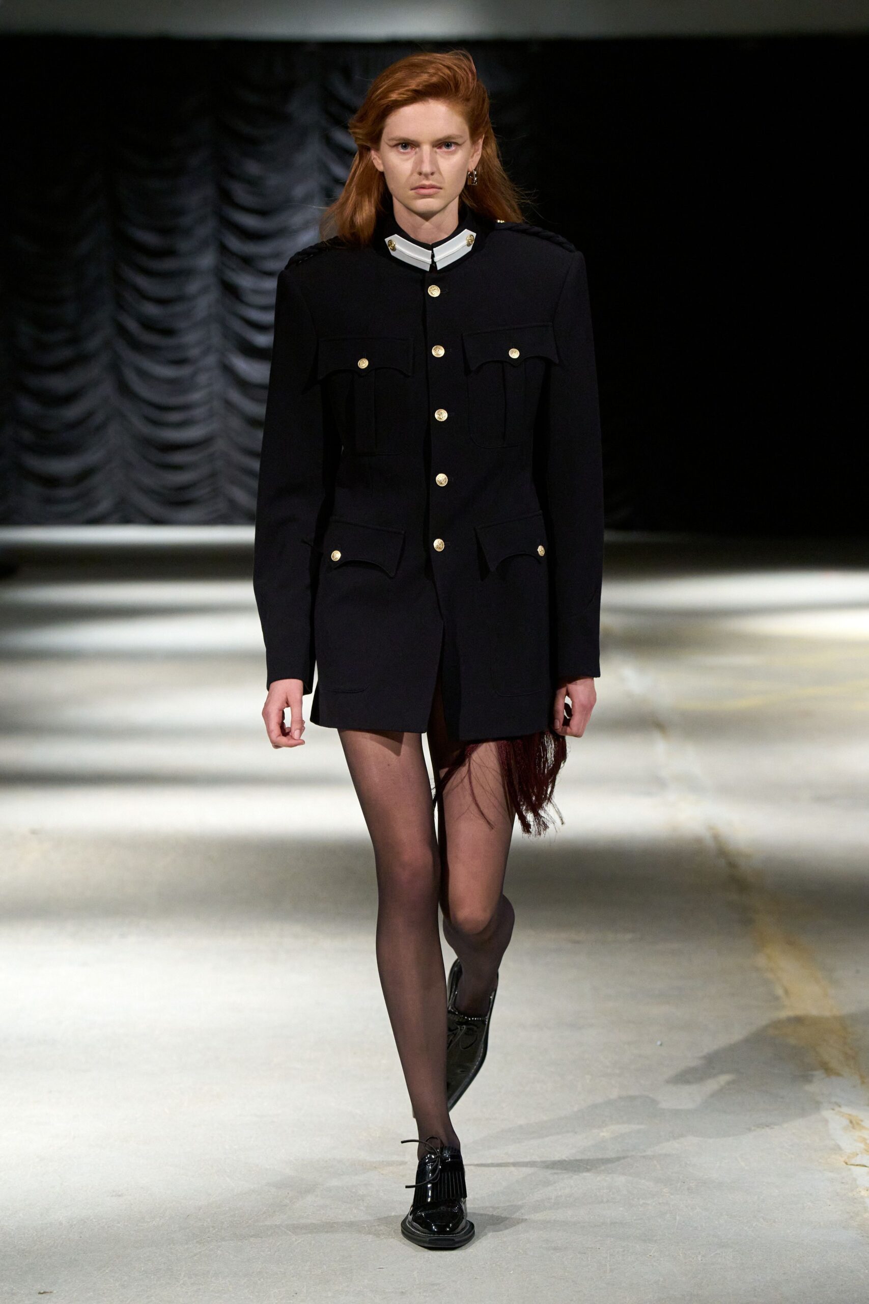 Black military coatdress with a white stand collar and gilt buttons. Burgundy fringe spilling from the right hem slit. Sheer smoke tights. Black patent loafers with kiltie fringe and thin bow laces. Single silver hoop. For crossing a hotel corridor after the ballroom emptied but the band still plays.
