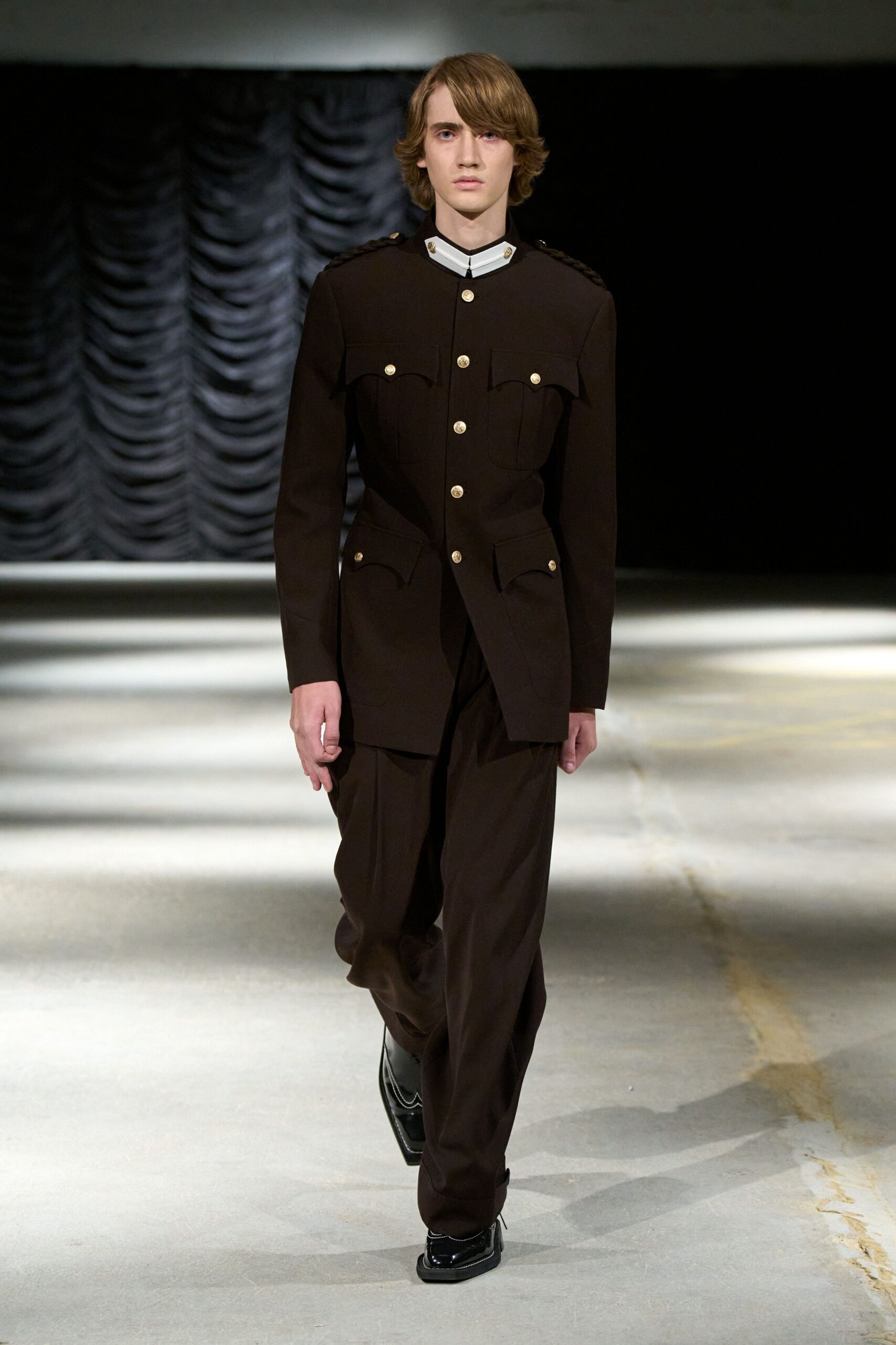 Dark brown tunic jacket with braided shoulder trims and bright crest buttons. Stiff white standing collar, split at the throat with tiny gold studs. Loose dark trousers breaking heavily over the shoe. Black patent square-toe derbies with white piping. Crossing a drafty civic hall before the school orchestra starts.