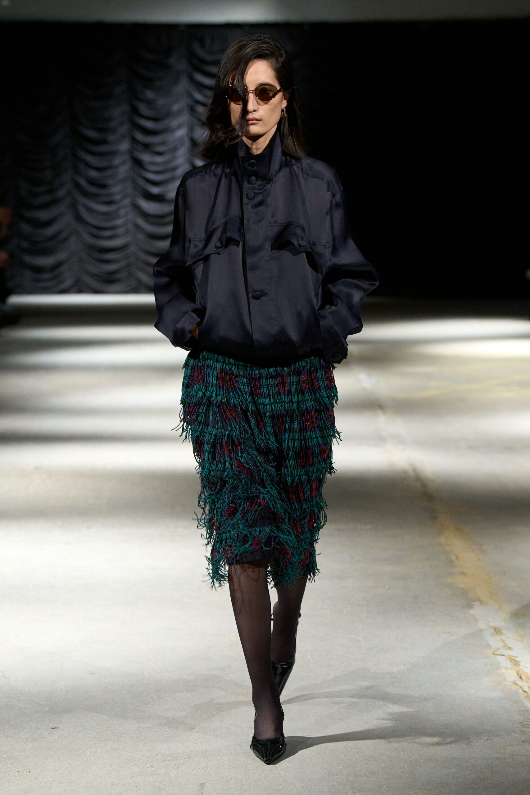 Amber oval lenses in a fine gold wire frame. Ink satin blouse with a high stand collar and covered buttons. Midi skirt crosshatched with teal fringe over a wine-and-navy plaid. Sheer black tights. Pointed black pumps with a narrow v-shaped throat. Walking the hotel corridor after the banquet, hem still catching chair splinters.