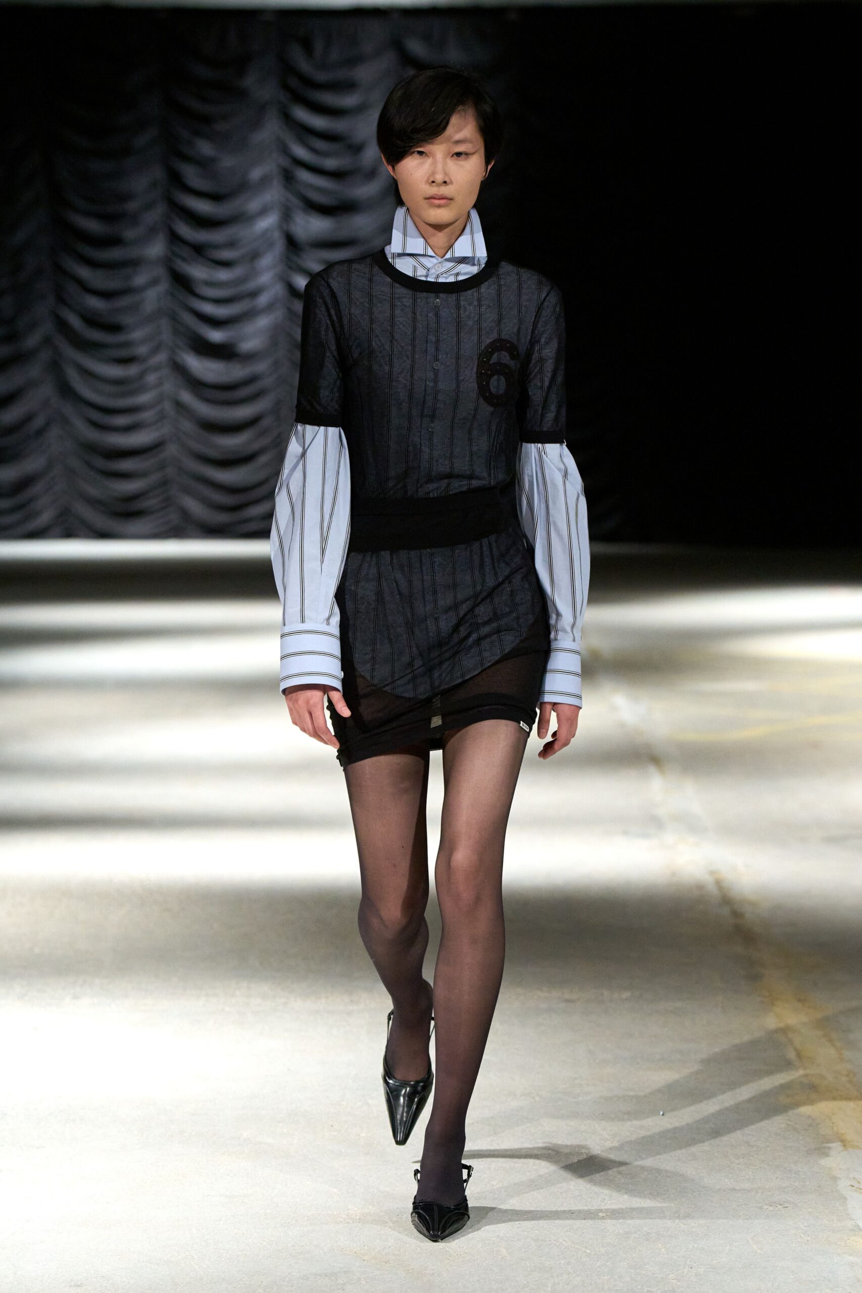 Pale blue striped shirt with long cuffs and a high spread collar. Black pinstriped mesh dress, ribbed trims and a sequined numeral six. Asymmetric black mini, one side hitched higher at the thigh. Smoke sheer tights. Black pointed pumps with a pinched vamp. Crossing the lobby carpet after an office party emptied into rain.