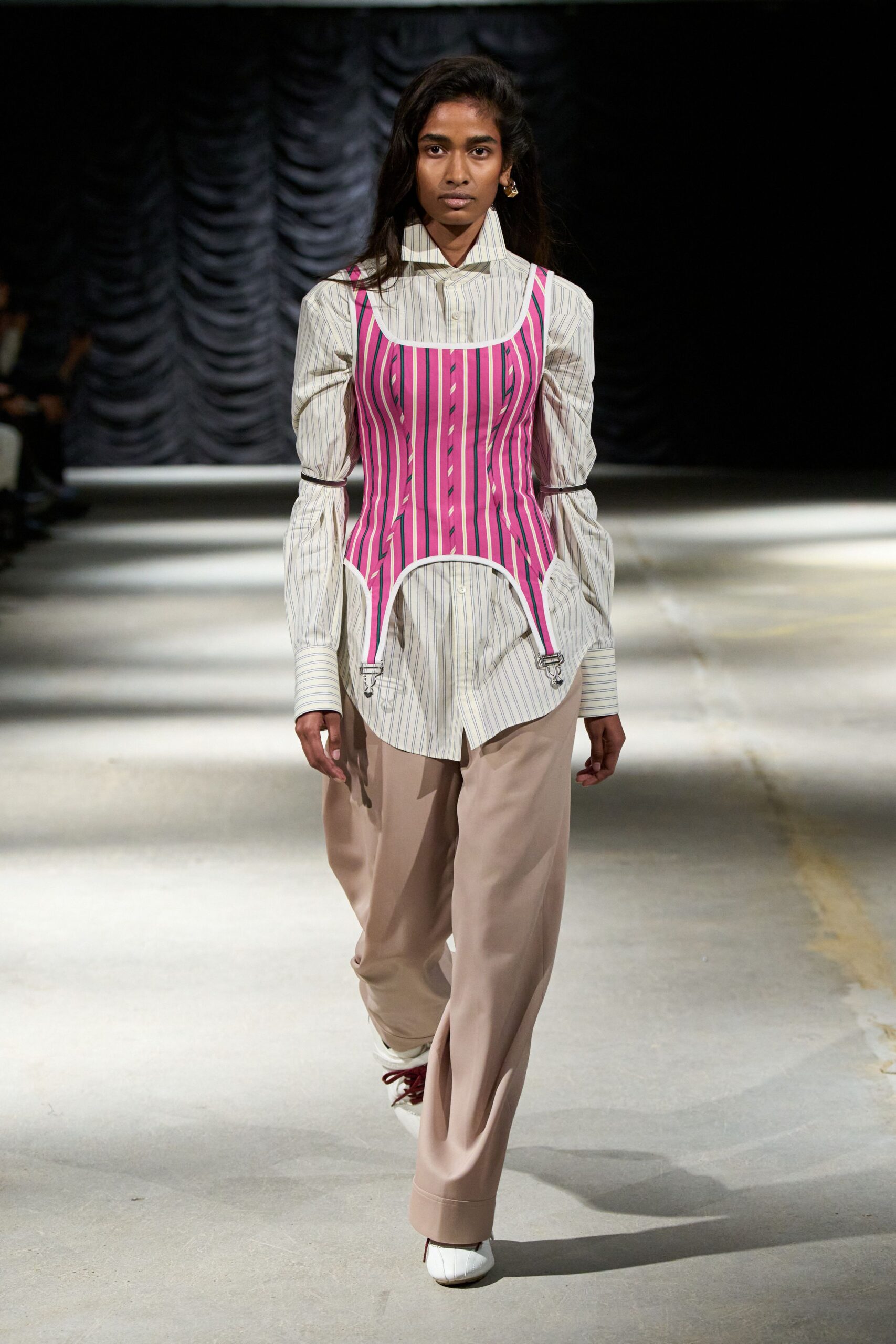 Cream striped shirting, collar flared high and cuffs stretched past the wrists. Hot-pink striped corset with white binding and suspender clips hanging loose. Blush wide-leg trousers breaking softly over the shoe. White square-toe shoes with oxblood laces. Walking to the registrar with your documents bent in a paper folder.
