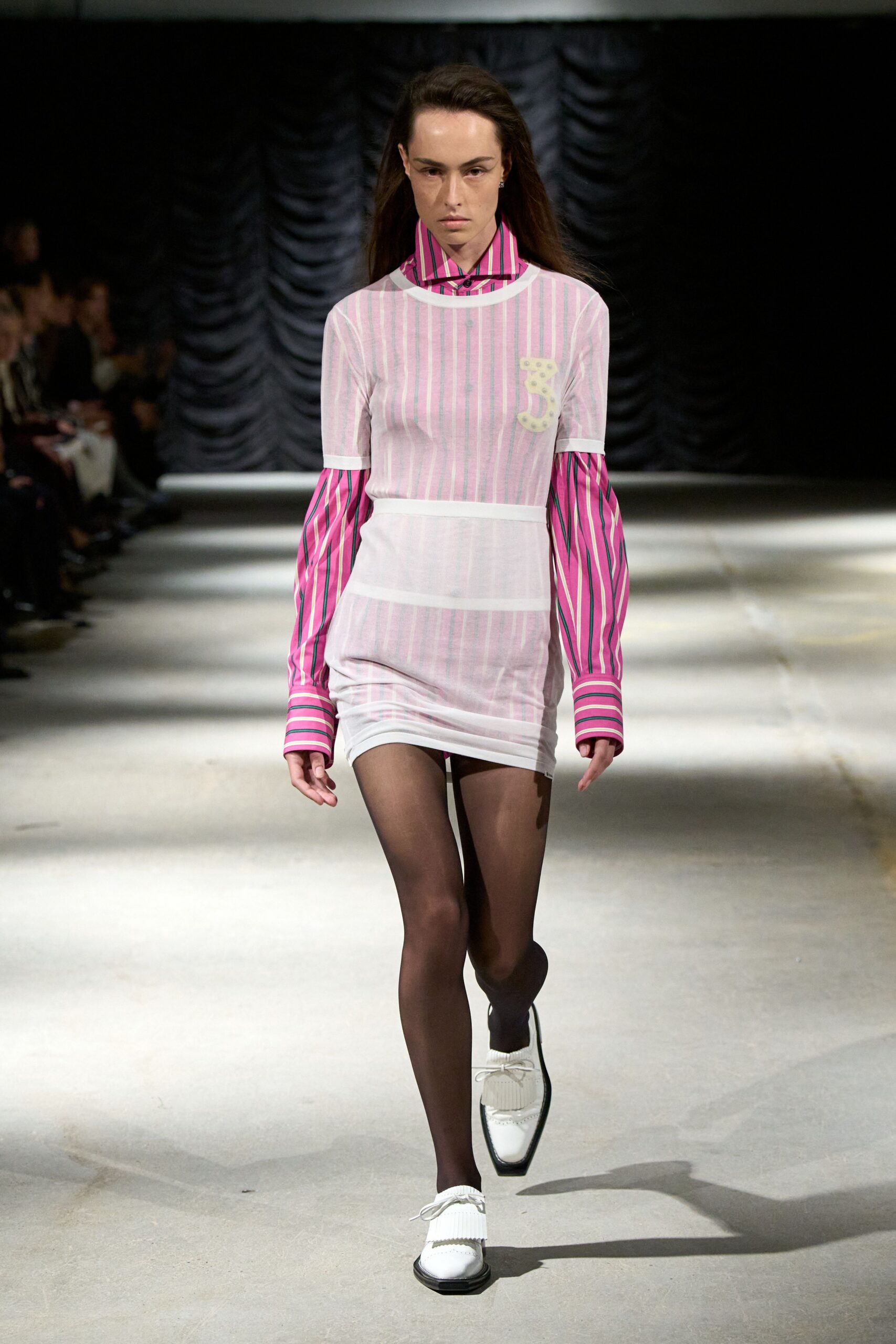 Hot-pink striped button-up with extra-long cuffs swallowing the hands. Sheer white mesh mini, pink pinstripes and a cream numeral 3 patch. Smoke-black sheer tights. White square-toe loafers with ribbed kiltie fringe and thin laces. Walking to the corner shop in school uniform after sleeping somewhere else.