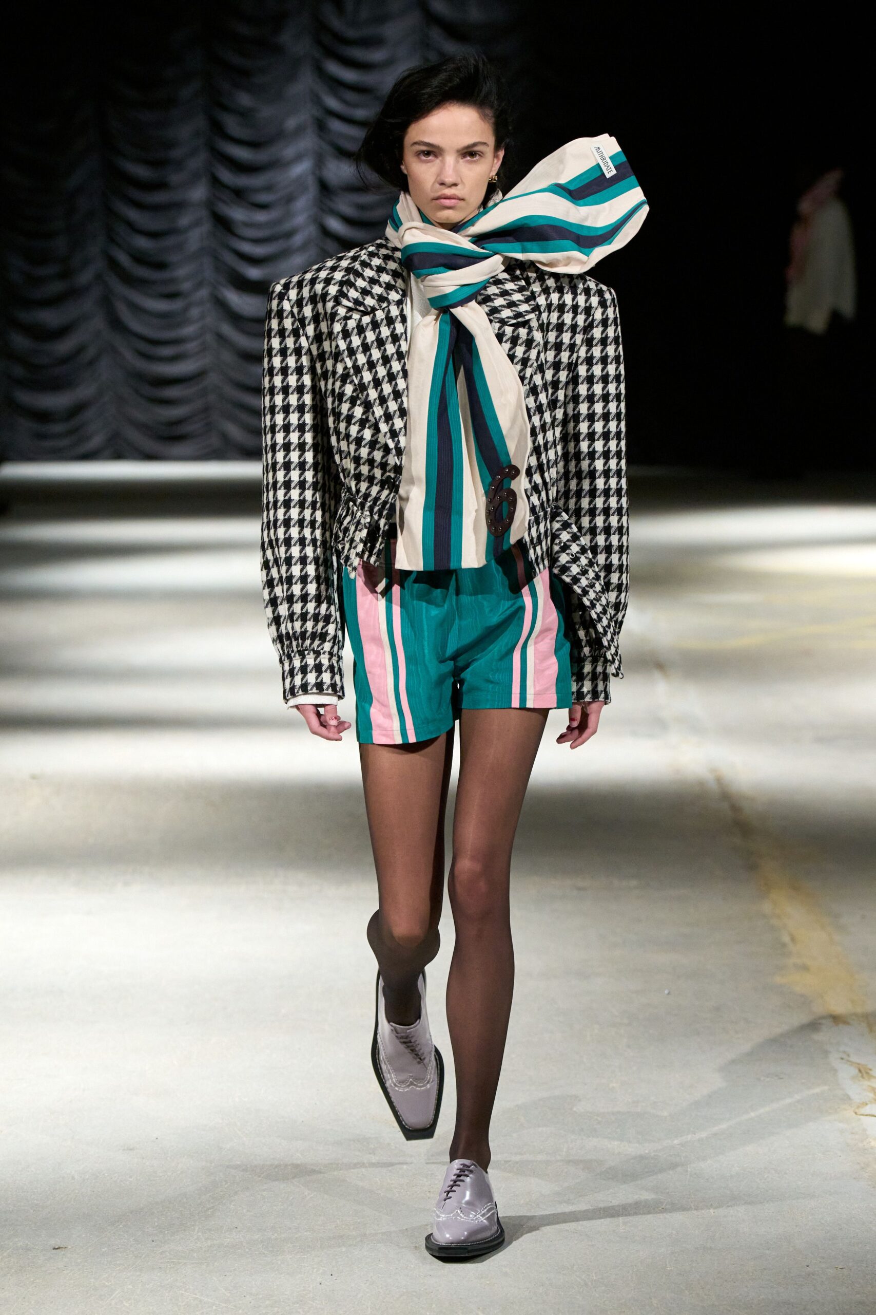 Oversized cream scarf in teal and navy stripes, blown into a stiff bow. Black-and-white houndstooth blazer, ruched in sharply at the waist. Teal shorts with pink side stripes and a papery sheen. Sheer black tights. Pale lilac patent oxfords with wingtip broguing. Crossing a windy pier after buying postcards you forgot to mail.