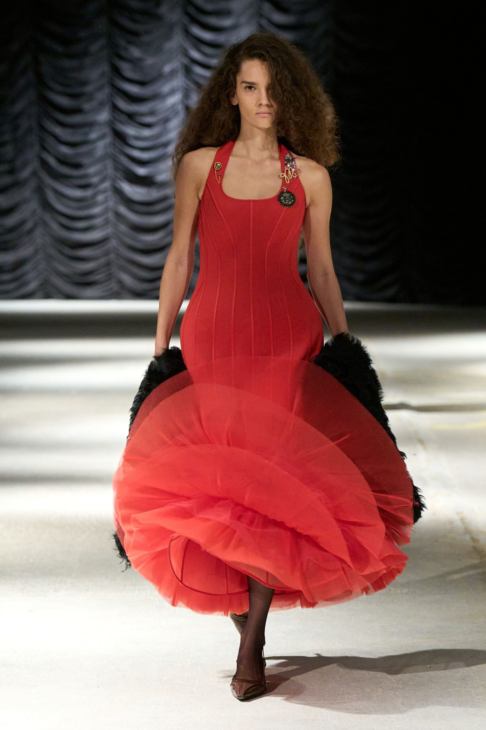 Tomato red knit halter dress, seamed through the torso into a huge wired tulle spiral. Mismatched pins clustered at the straps, one dangling on a short chain. Black shaggy fur caught behind the hips. Sheer smoke tights. Brown pointed pumps. Outside the opera house after midnight, still carrying your own applause.