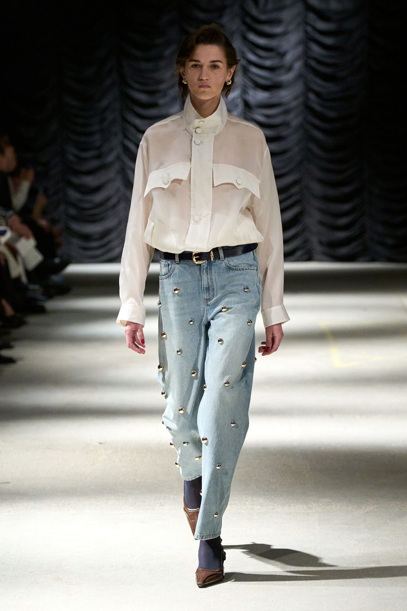 Sheer ivory blouse with a tall stand collar and curved flap seams. Narrow black belt with a small gilt buckle. Pale denim jeans scattered with domed metal studs, one climbing the fly. Opaque navy tights. Brown square-toe pumps with a raised center seam. Walking into a civic theater matinee after missing the dress code memo.
