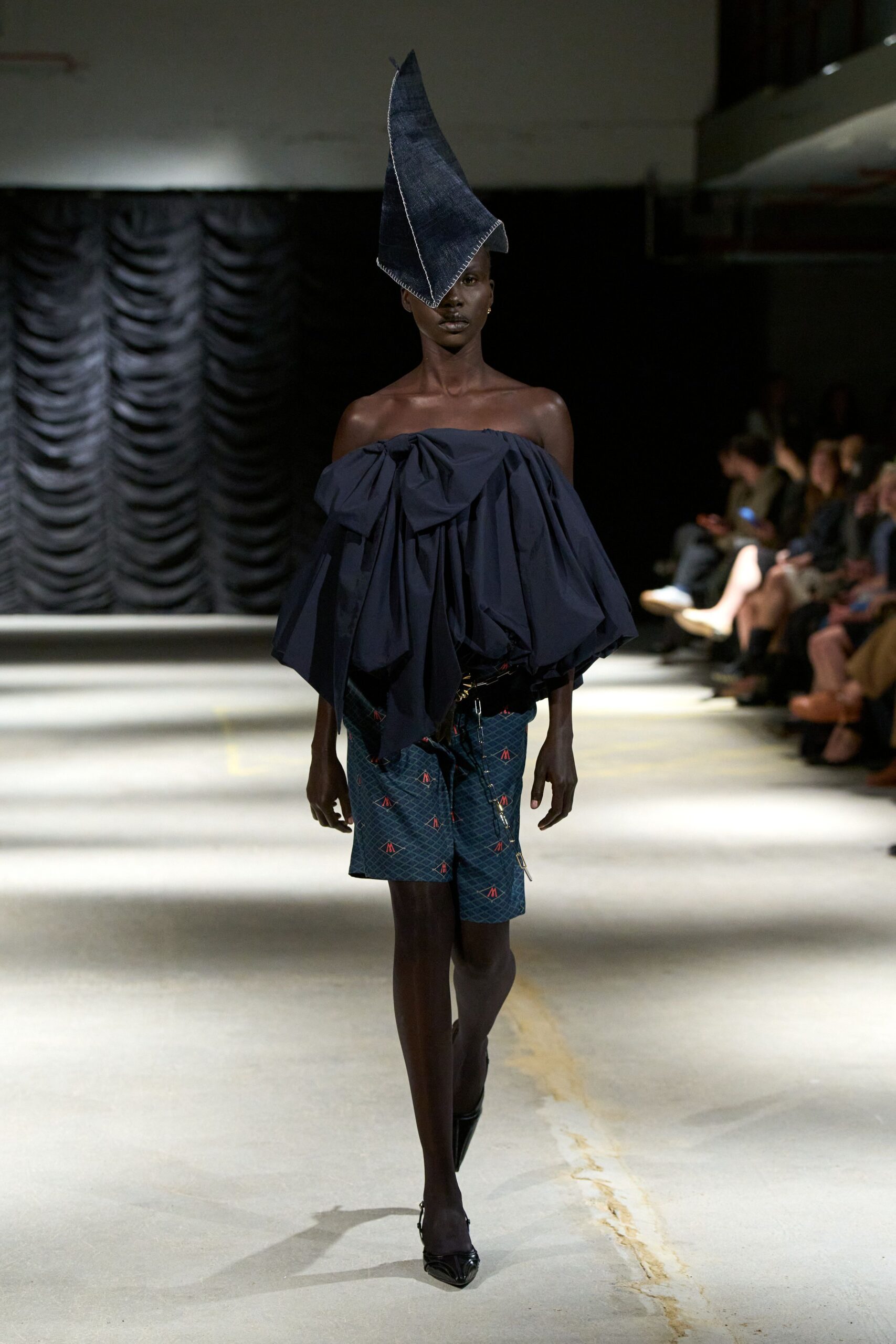 Indigo denim cone hat with blanket-stitch edges, tipped over one eye. Off-shoulder navy cotton top gathered into dense balloon folds. Teal monogram shorts with a slanted zip and glossy black waistband. Black patent pointed pumps. Crossing a drafty civic hall after midnight with the raffle money.