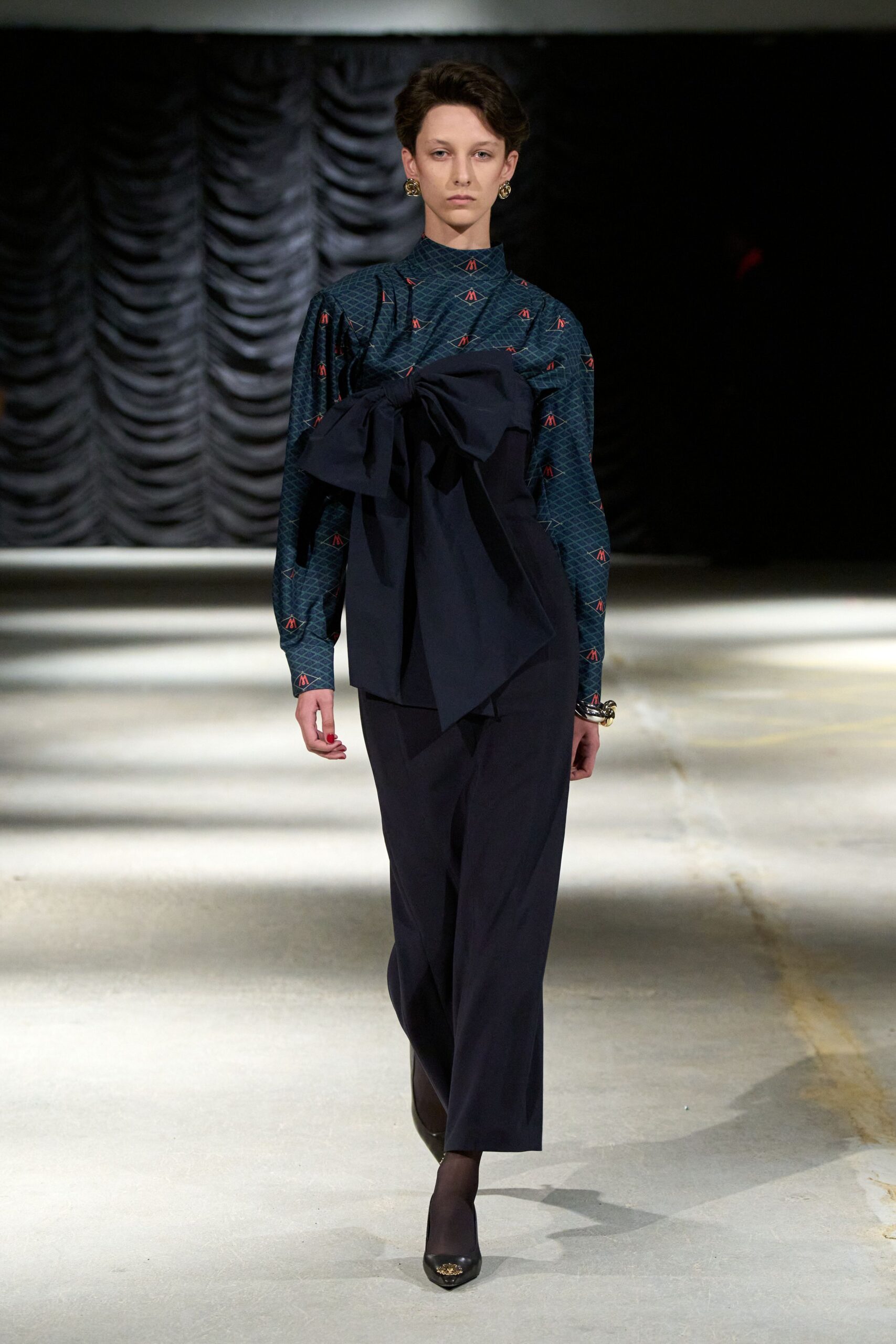 Teal mock-neck top in fine diamond print with tiny red motifs. Navy cotton bow panel wrapped across the chest, one tail hanging long. Loose navy trousers with a soft front crease. Chunky gilt cuff. Black pointed pumps with a small gold toe ornament. Matinee crowd thinning out under theater drapes and weak winter sun.