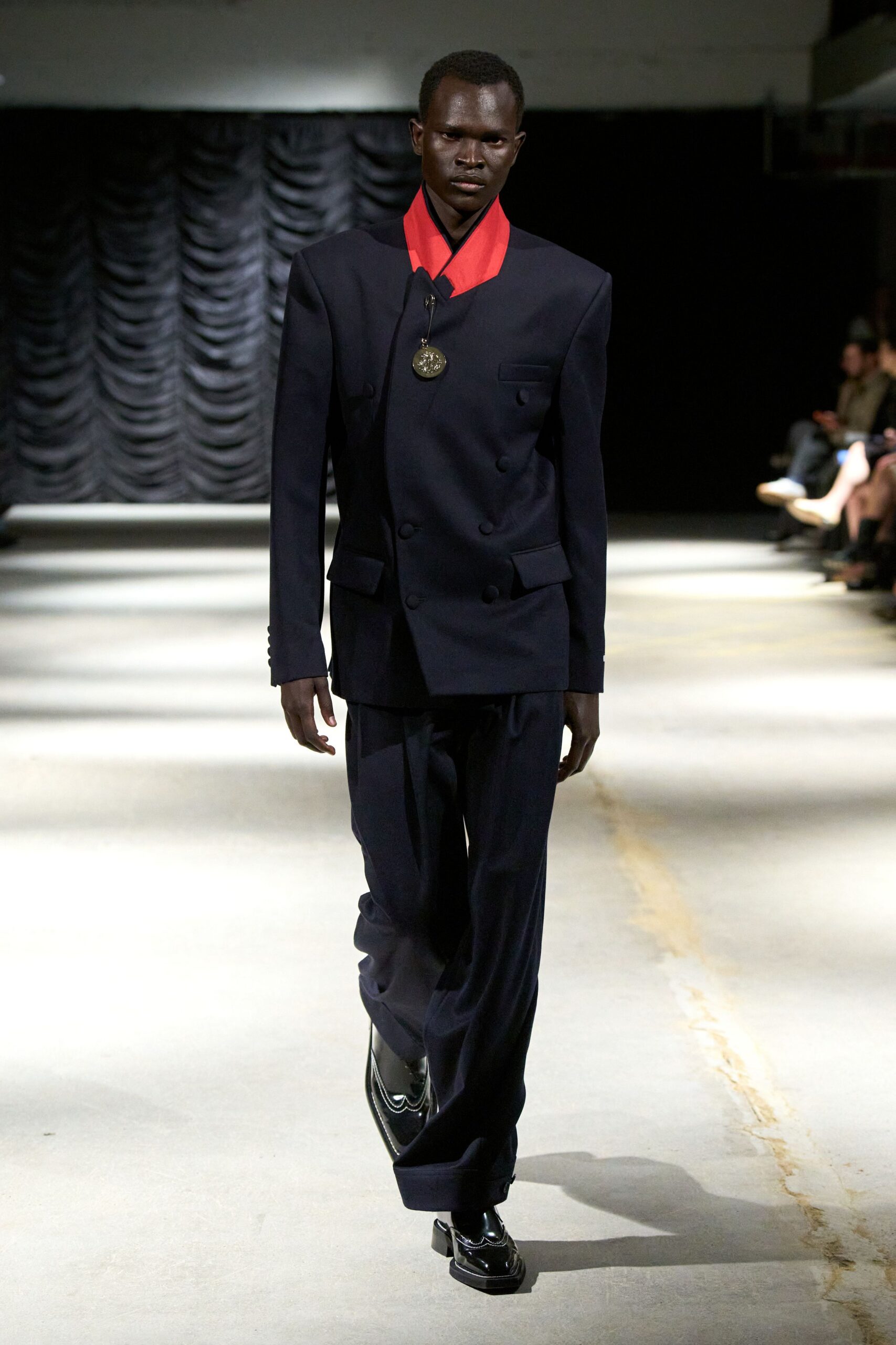 Ink wool double-breasted jacket with a vivid red shawl collar. Round metal medallion hanging from a narrow black cord. Loose dark trousers breaking heavily over the shoe. Black patent shoes with white piping tracing the apron seam. For crossing a hotel lobby after midnight with a medal you did not earn.