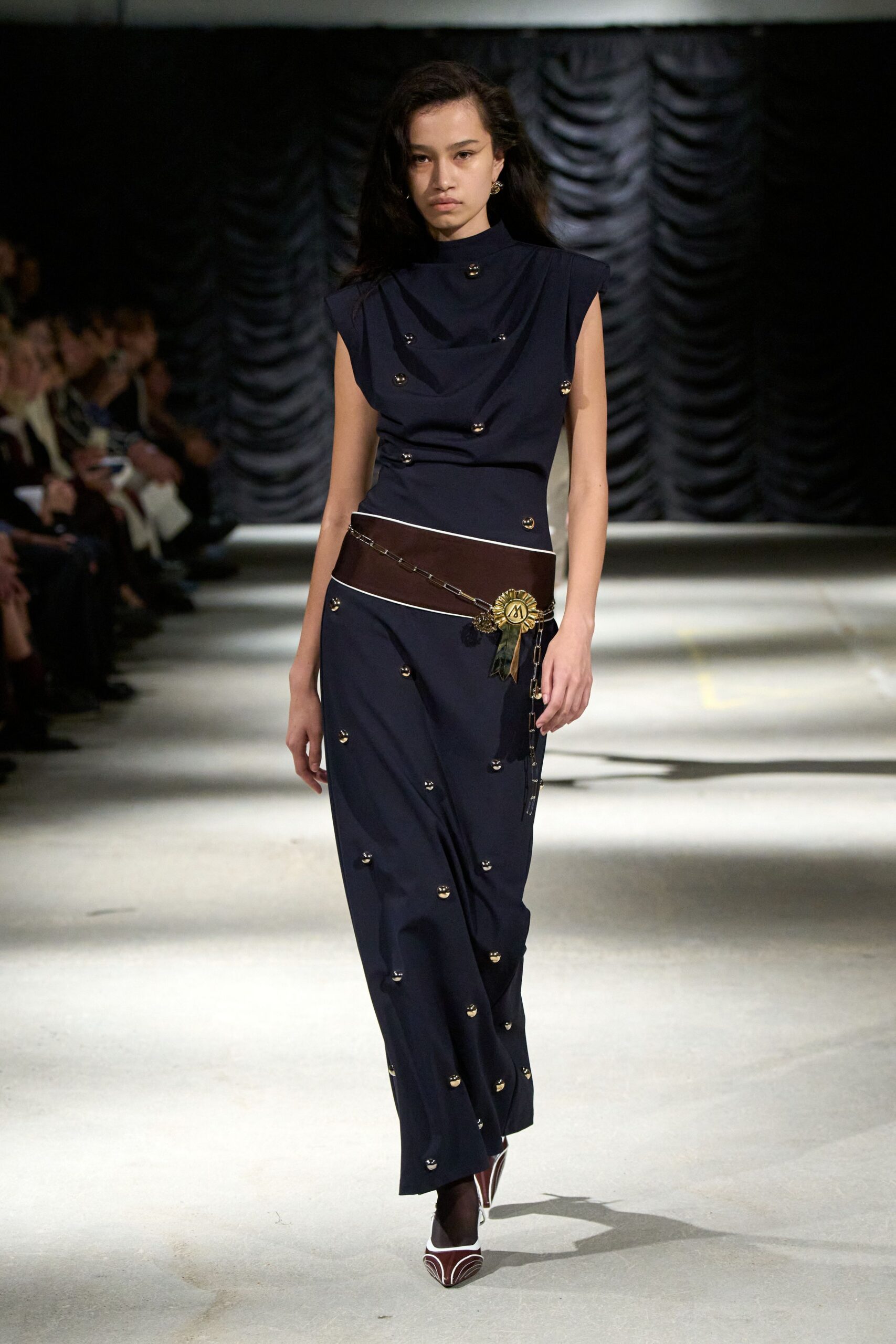 Navy sleeveless column dress with a draped bib front and scattered dome studs. Wide oxblood leather hip belt, pinned with a rosette and trailing chain. Sheer black tights. Oxblood pumps traced with white piping at the pointed toe. For collecting a prize at the town hall after dark.