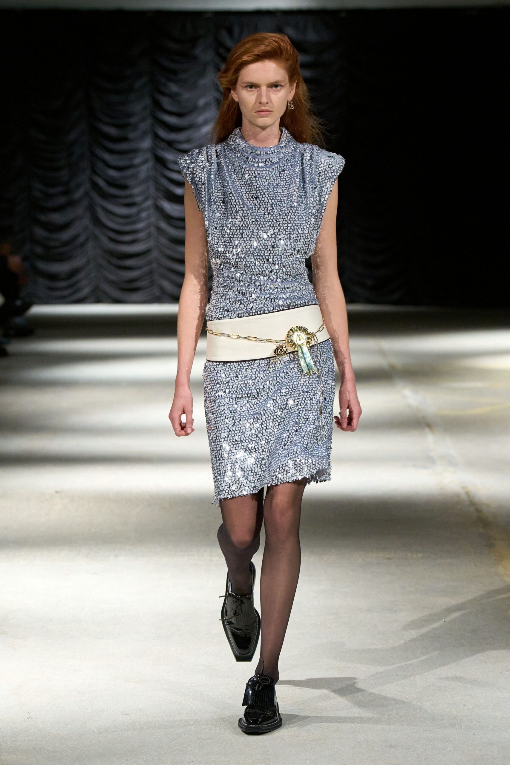 Ice-blue sleeveless dress crusted in tiny paillettes, shoulders pushed into soft points. Wide white leather belt with a rosette medal and fine chain drape. Sheer smoke tights. Black patent loafers with long fringe over the vamp. For collecting a prize at the village hall after rain.