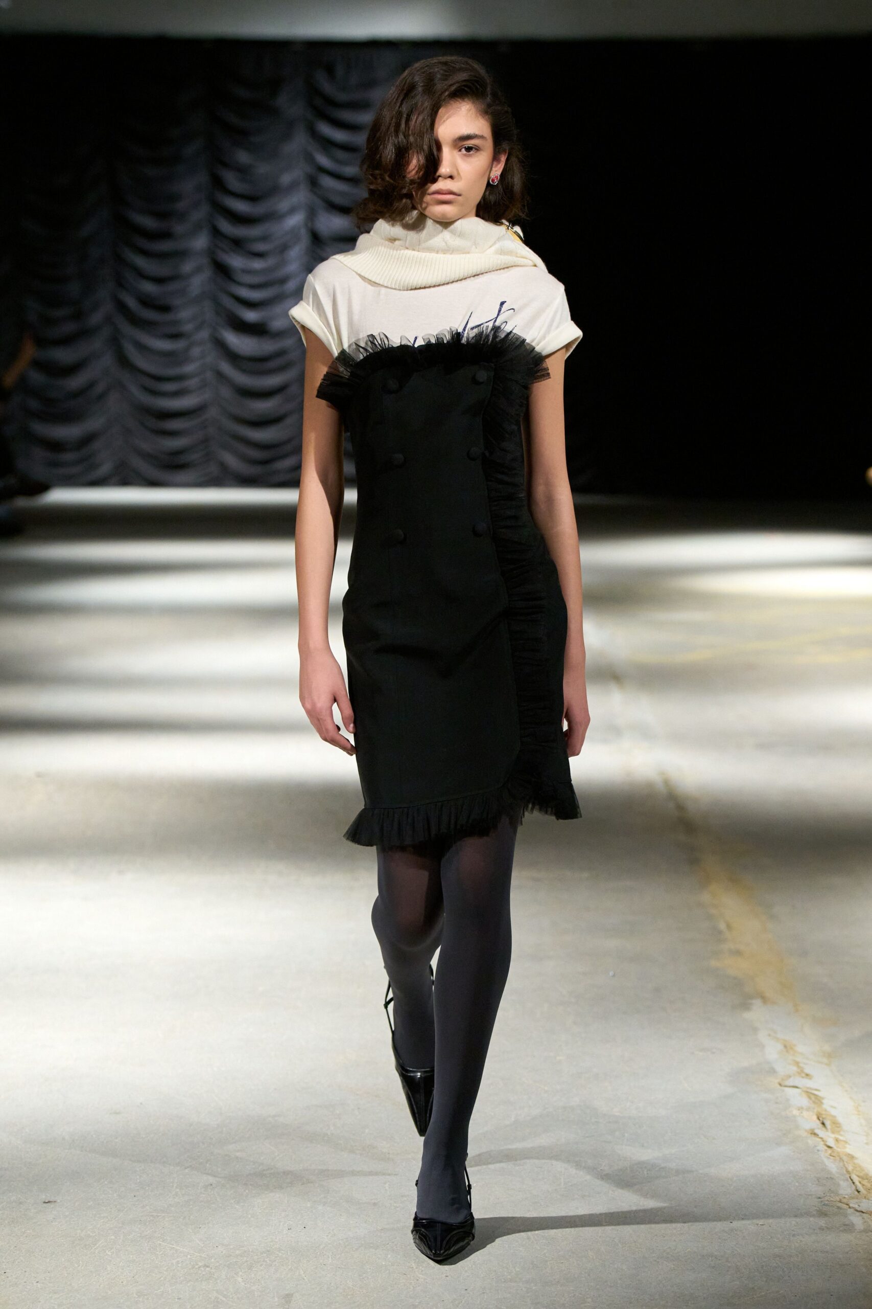 Cream ribbed knit looped high, collapsing into a soft cowl. Black strapless dress with tulle ruffle crest and fabric-covered buttons. Smoke-black opaque tights. Patent black pumps with razor-point toes. Matinee crowd thinning, still carrying theatre dust on your sleeves.