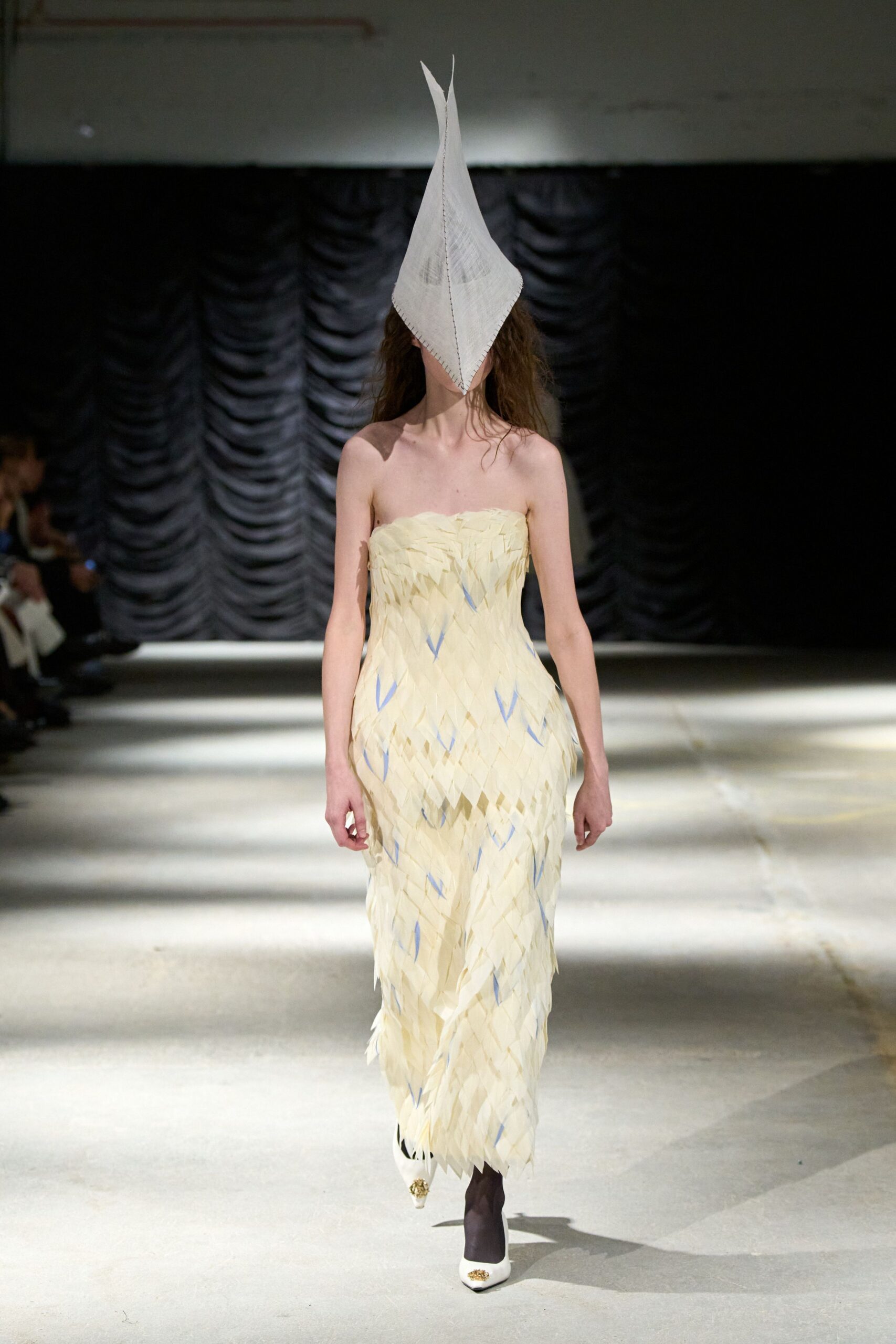 Tall white pointed cloth mask, blanket stitching running down the center. Strapless ivory column dress built from layered petal-like strips, flecked with pale blue. Dark sheer tights. White pointed pumps with gilt toe ornaments. Crossing a drafty town hall after the pageant has gone strange.