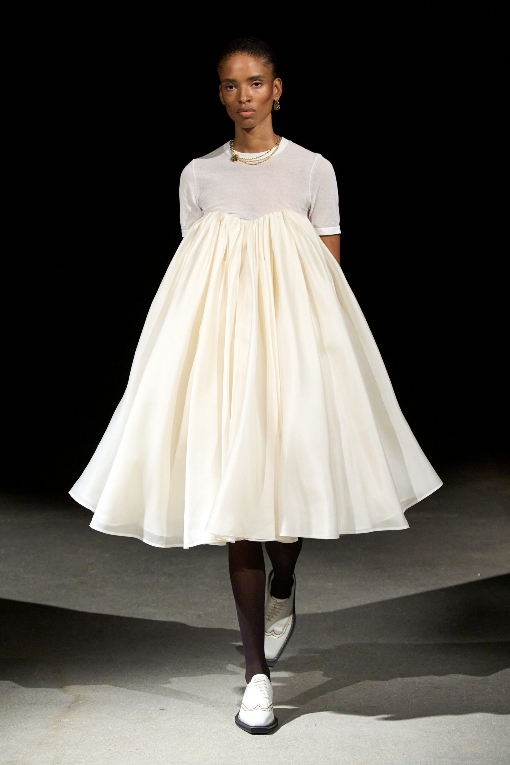 Double gold chain with a knotted cluster off center. Ivory baby-doll dress, sheer knit yoke over a gathered sateen skirt. Dark brown sheer tights. White leather oxfords with brogue scallops at the square toe. Sunday recital, coat check ticket still tucked in your palm.