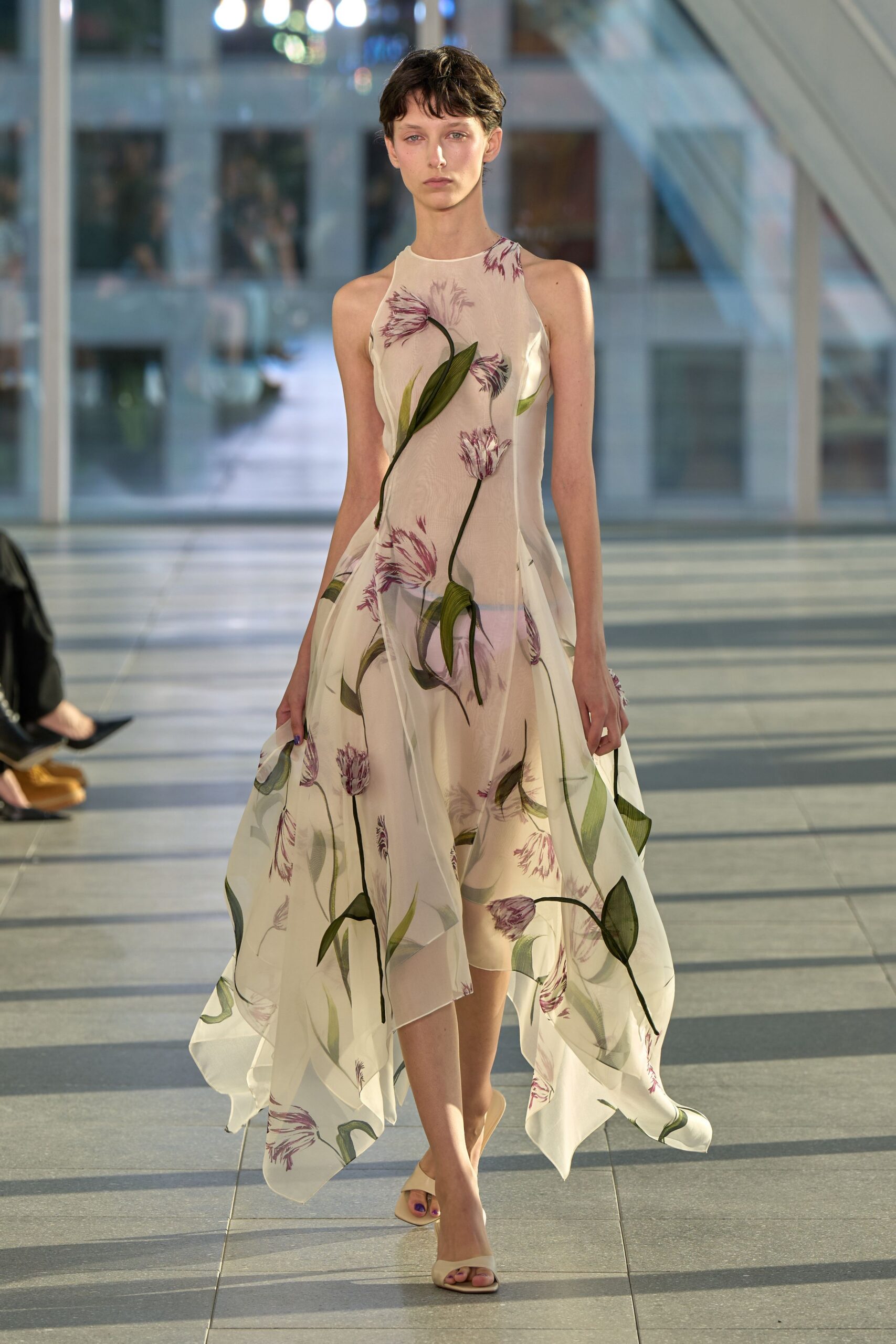 Sheer ivory organza dress printed with mauve tulips, handkerchief hem lifting at every point. Pale blush underlayer showing cleanly through the gauzy skirt. Barely there cream leather slide sandals. Crossing a glass atrium just after the florist has finished for the day.