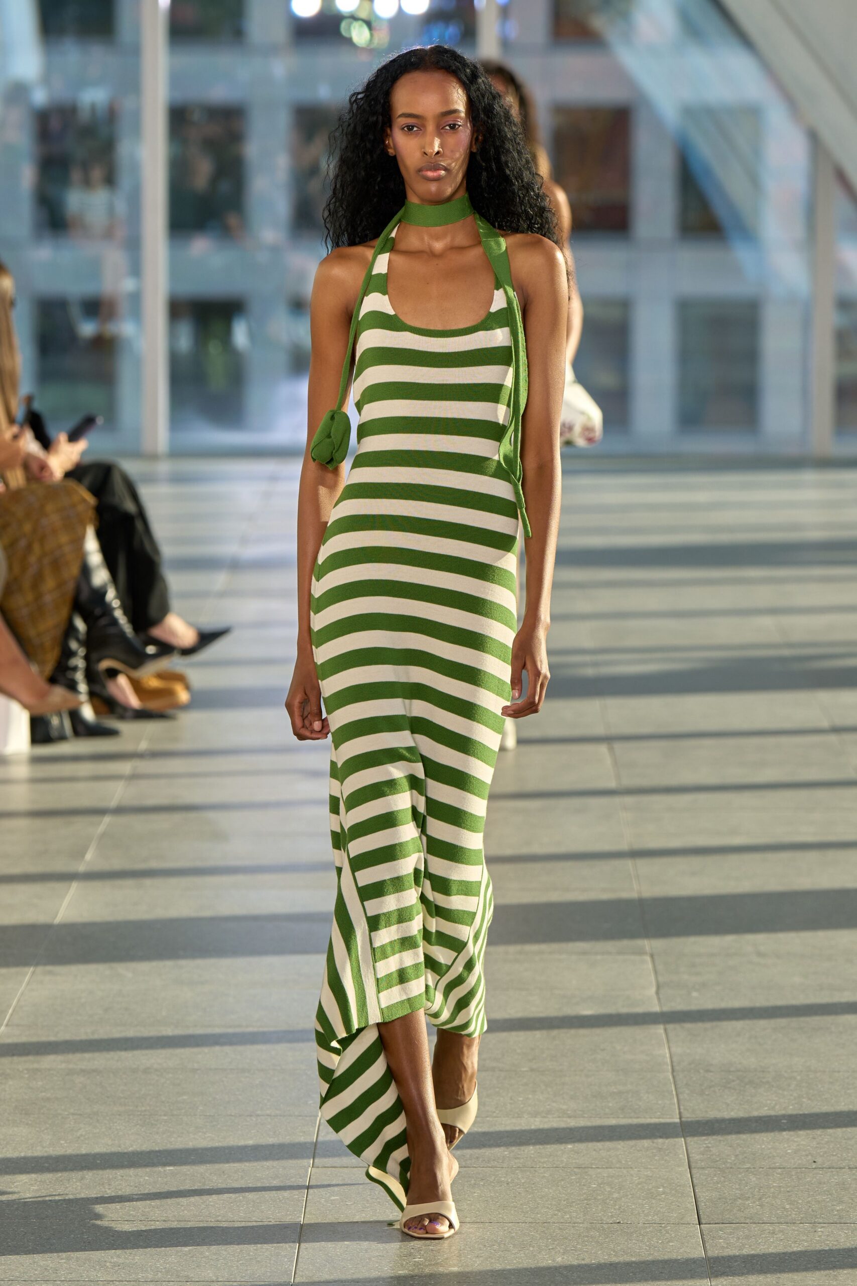 Green-and-cream striped knit halter dress, hem dipping longer behind. Wide green band circling the throat, ties trailing down. Tiny green pouch hanging from a narrow shoulder cord. Cream slip-on mules with a squared open toe. Walking the hotel atrium after a poolside lunch ran pleasantly long.