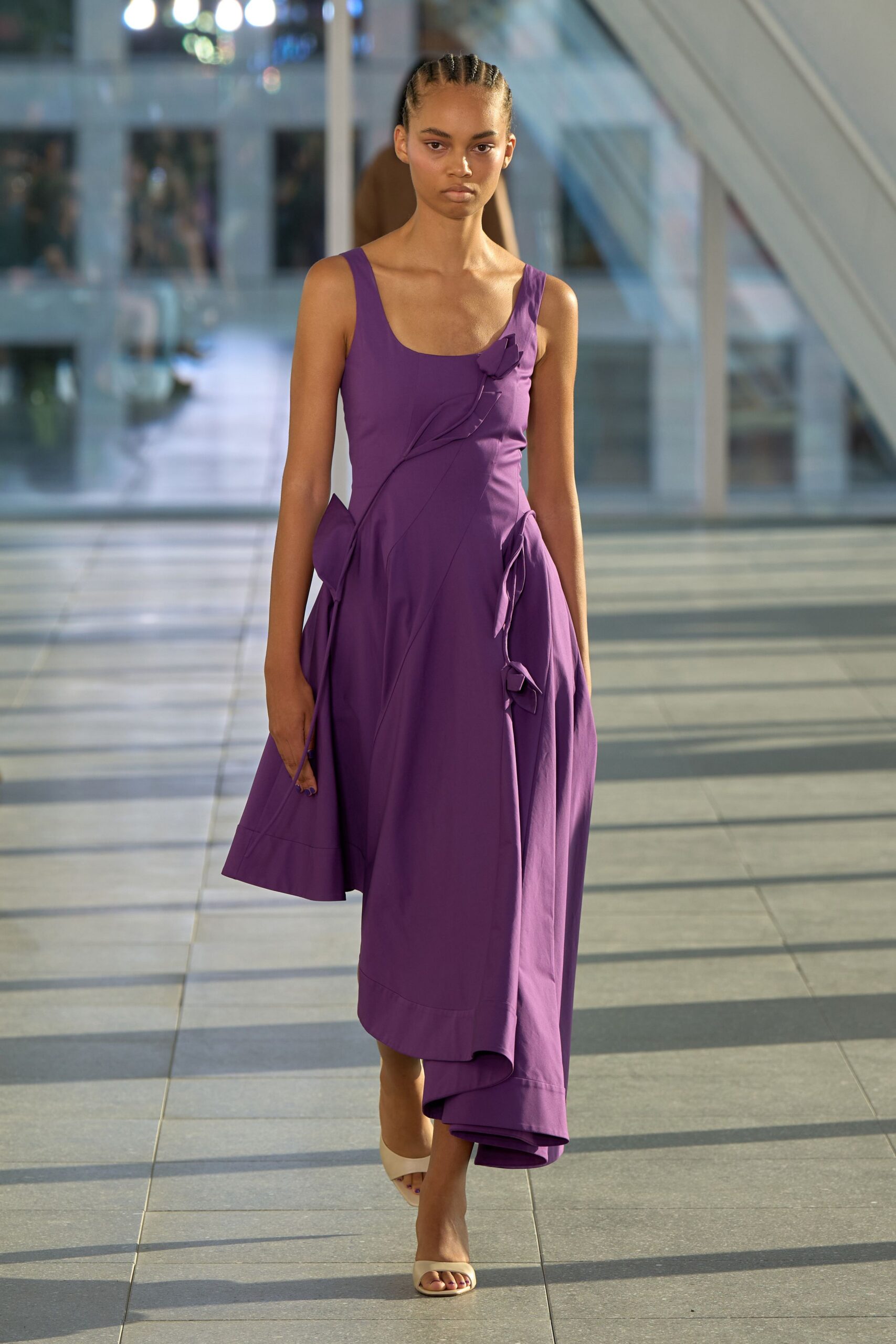 Violet scoop-neck dress with spiraling seams and petal flares at the hips. Cream leather slide mules, square at the open toe. Crossing a glass concourse in late sun, carrying nothing but your key card.
