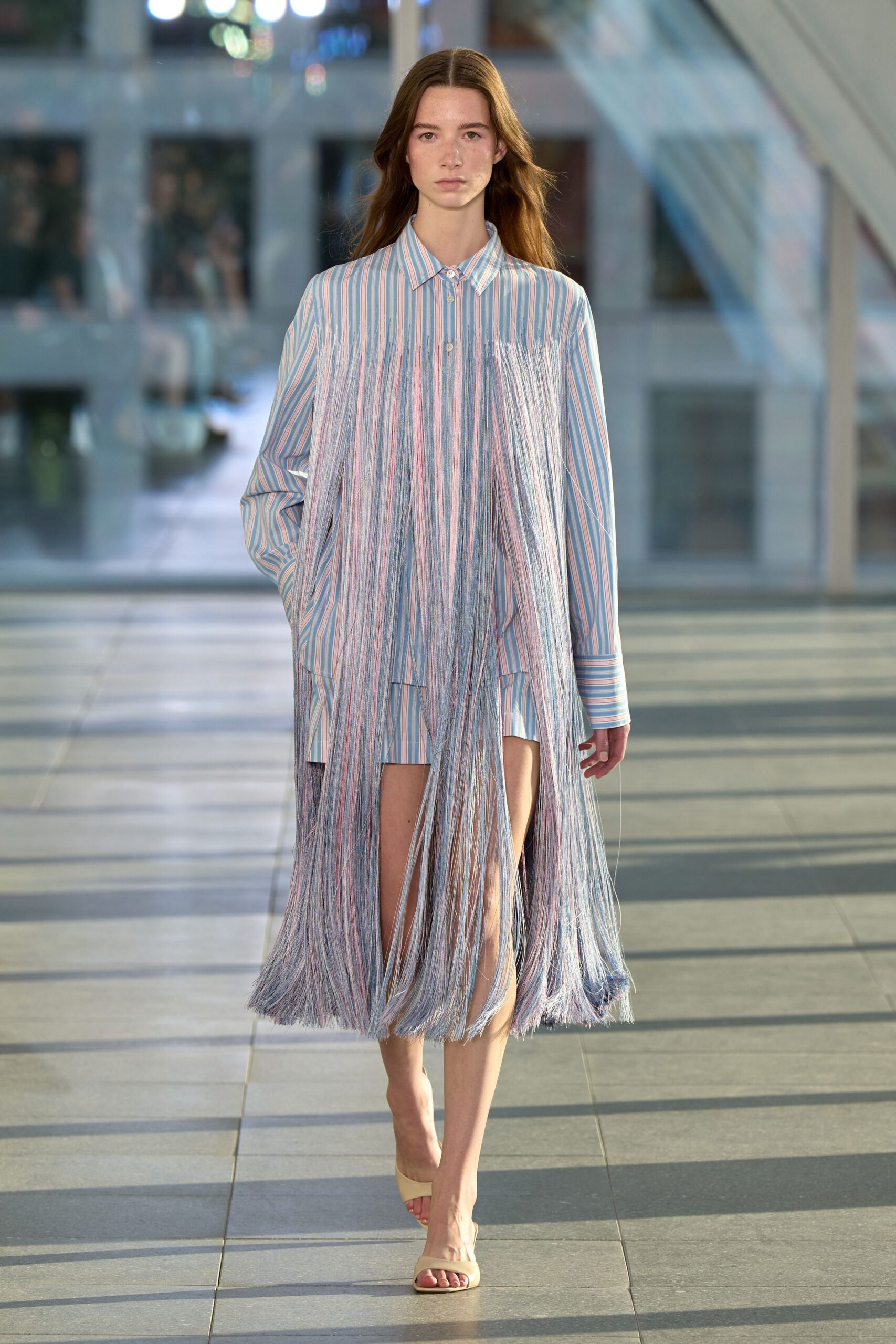 Pale blue and pink striped shirting, buried under curtain-length fringe from the yoke. Cream leather slide mules with a squared toe. Crossing a glass atrium when the sun starts striping the floor.