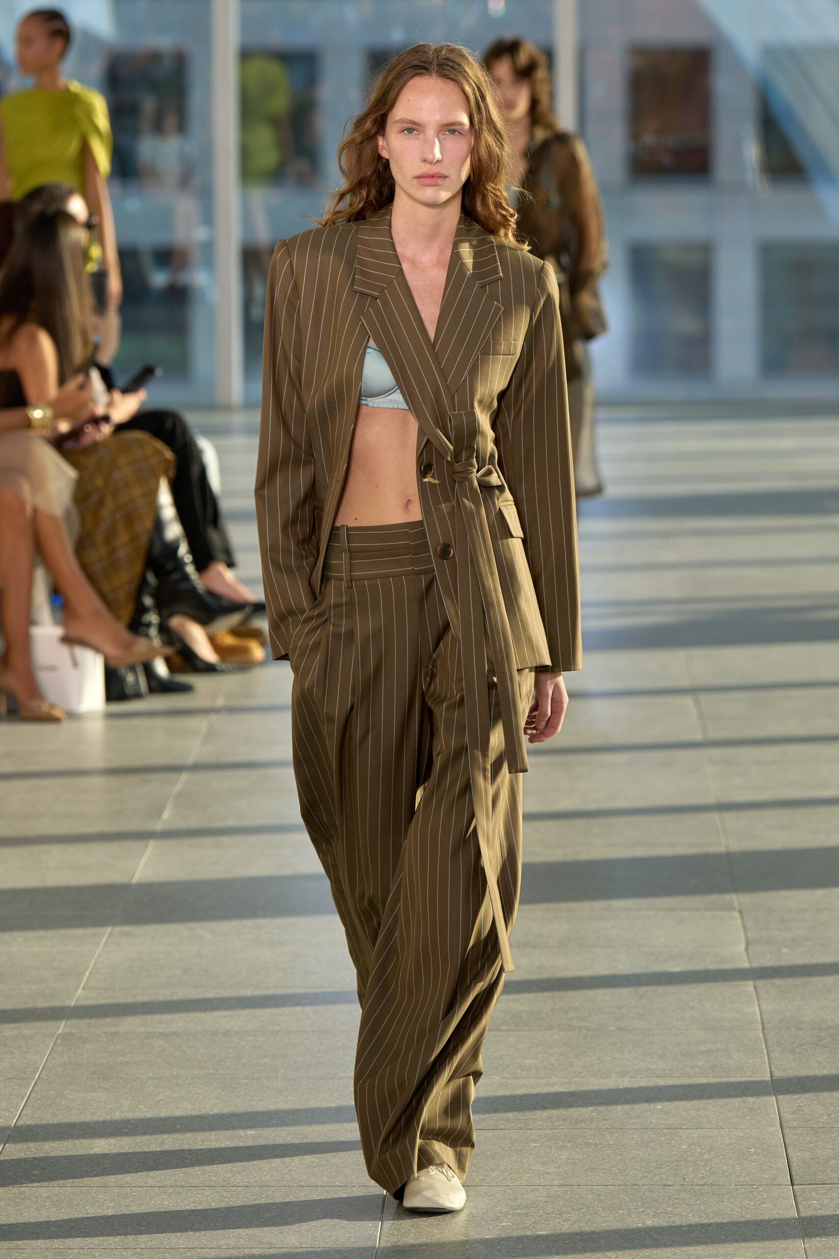 Olive pinstripe blazer wrapped with a self-tie knot through the waist. Pale blue satin bralette peeking from the deep crossover opening. Loose pinstripe trousers with a broad waistband and long pooled break. Cream square-toe boots. Crossing a glass atrium before the offices have fully filled.