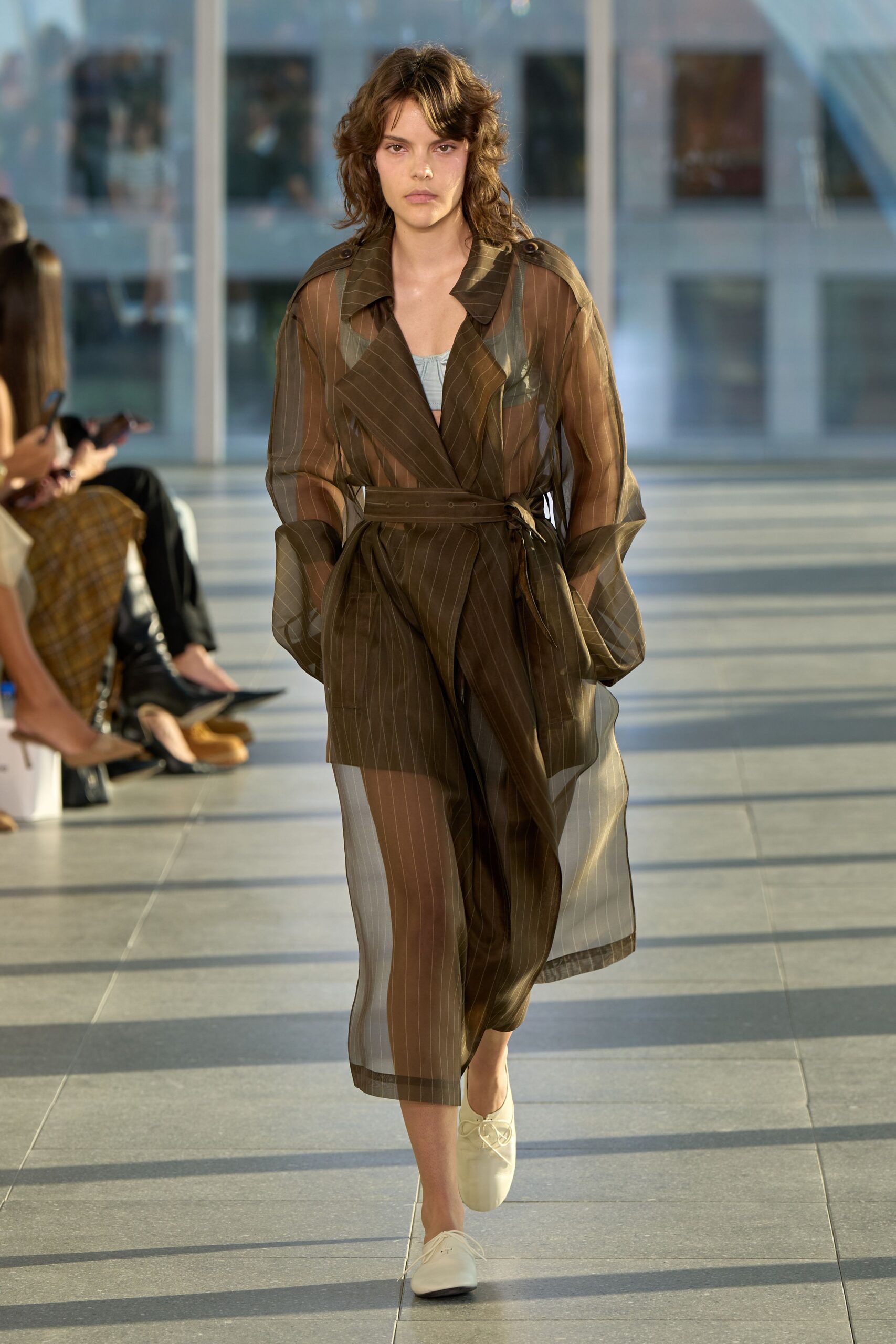 Sheer brown pinstriped trench, belt tied through a tiny side loop. Pale blue gathered bralette showing beneath the translucent lapels. Matching pinstriped shorts, cut high under the gauzy layers. Cream lace-up flats with blunt squared toes. Crossing an office atrium after rain, still carrying the heat outside.