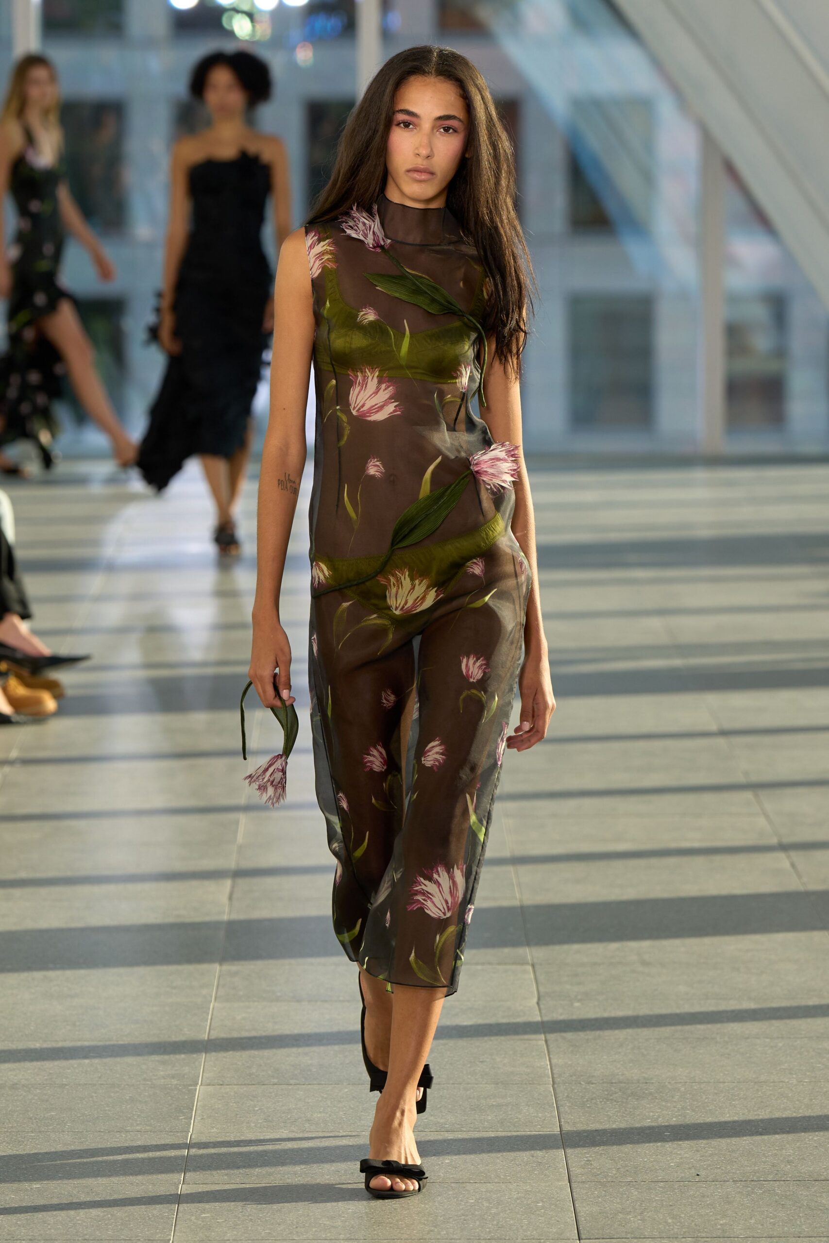 Smoke-brown mesh column printed with pink thistle blooms and trailing leaves. Olive briefs visible beneath the transparent layer. Black thong sandals with a knotted strap. Single drooping bloom carried loose in one hand. Crossing a glass atrium just before the florist shutters come down.